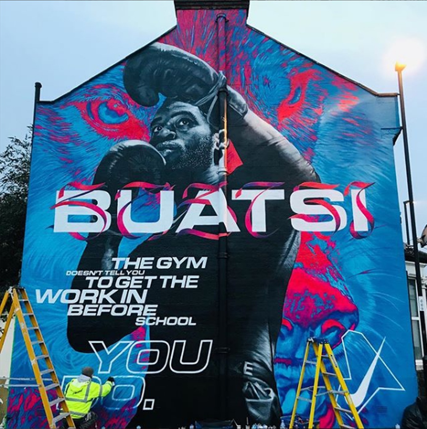 Duży mural na ścianie budynku przedstawiający boksera w rękawicach, z napisem 'BUATSI' i tekstem motywacyjnym, malowany przez artystę stojącego na drabinie w żółtej kurtce. W tle widoczne niebo...