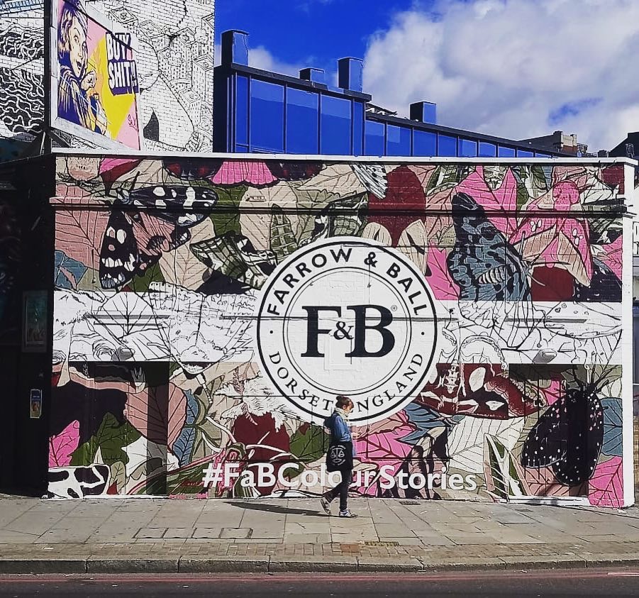 Duży mural na ścianie budynku z motywem liści i motyli w odcieniach różu, zieleni i szarości, z logo Farrow & Ball w centrum. Przechodząca osoba w maseczce dodaje skali. Nad muralem widać fragmenty...
