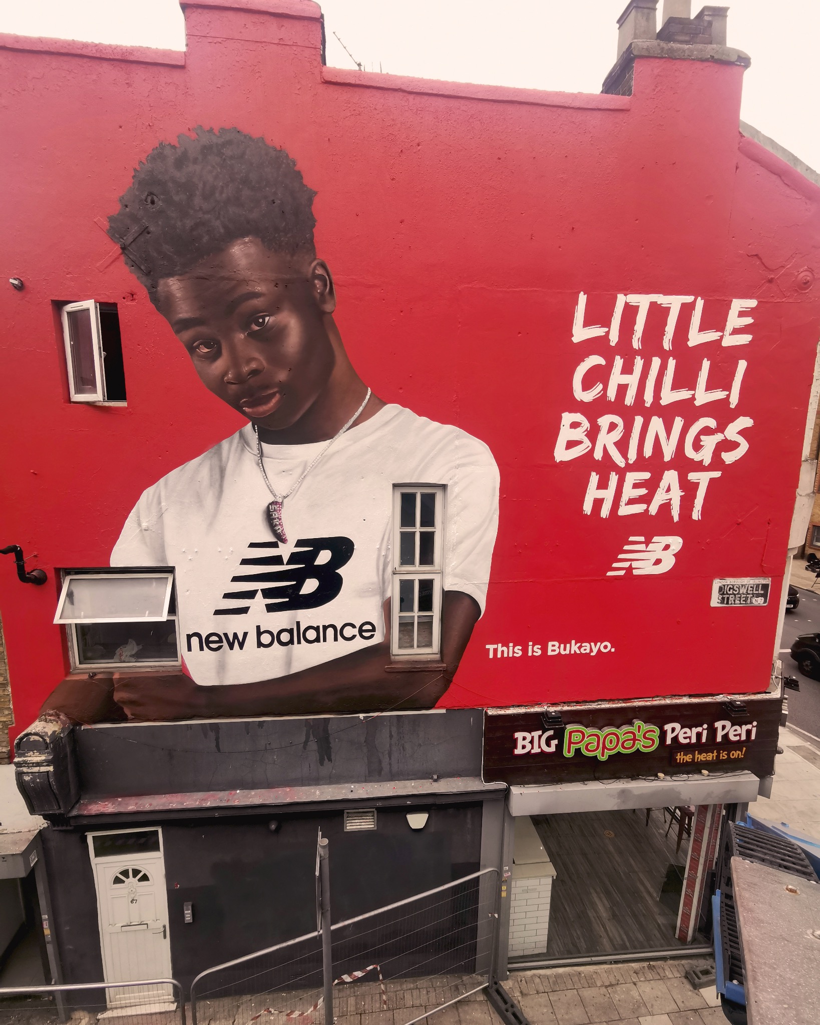 Duży mural reklamowy na czerwonej ścianie budynku, przedstawiający młodego mężczyznę w koszulce New Balance, z napisem 'Little Chilli Brings Heat'. Okna budynku wkomponowane w mural.