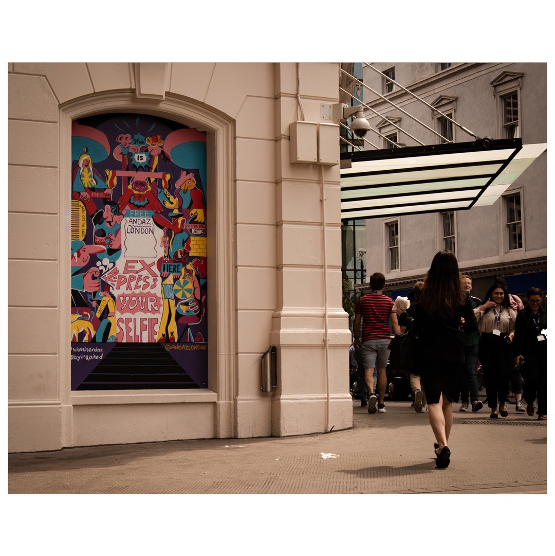 Kolorowa, abstrakcyjna grafika na ścianie budynku w Londynie, zachęcająca do zrobienia selfie, z przechodzącymi obok ludźmi.