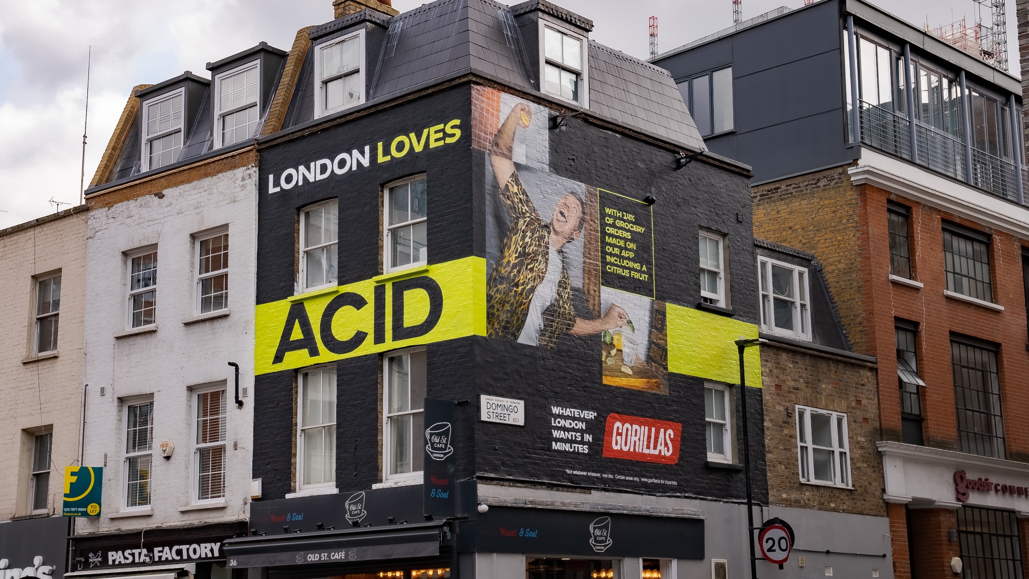 Czarna ściana budynku w Londynie z muralem reklamowym przedstawiającym osobę wyciskającą cytrynę, hasłem 'London Loves Acid' i logo firmy Gorillas, widoczne są także sąsiednie budynki o różnej...