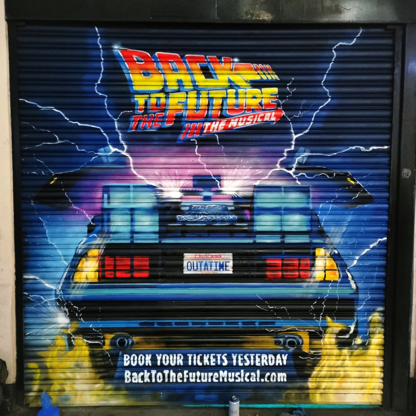 Mural na metalowej roletce sklepowej przedstawiający tył samochodu DeLorean z filmu 'Powrót do przyszłości' i tytuł musicalu, z efektem błyskawic i napisem 'OUTATIME' na tablicy rejestracyjnej.