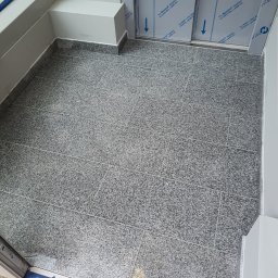 KRONOS - Wykończona podłoga w małym pomieszczeniu wyłożona szarymi, nakrapianymi płytkami, widok z góry na ułożone płytki i fragment progu.