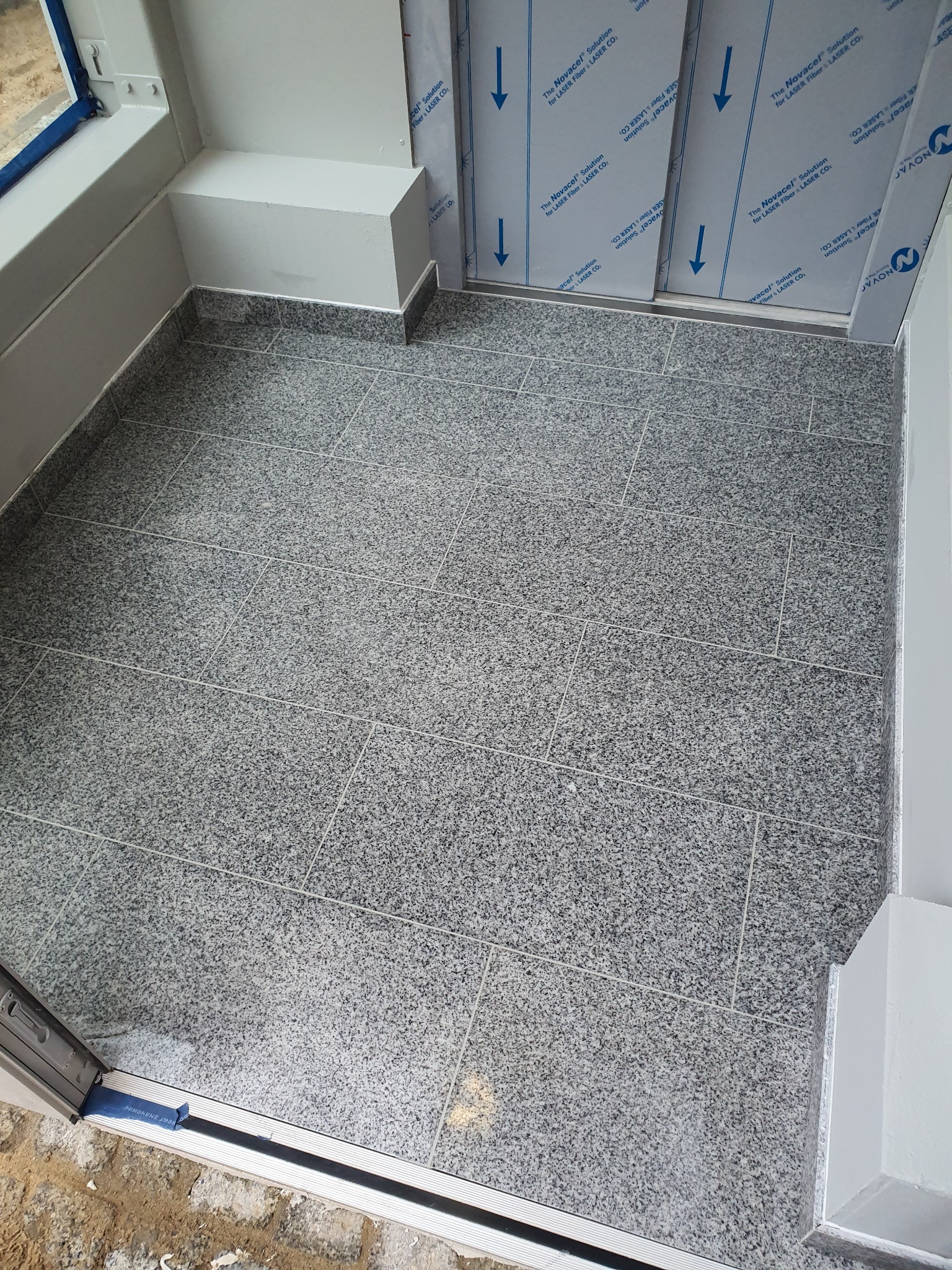 Wykończona podłoga w małym pomieszczeniu wyłożona szarymi, nakrapianymi płytkami, widok z góry na ułożone płytki i fragment progu.