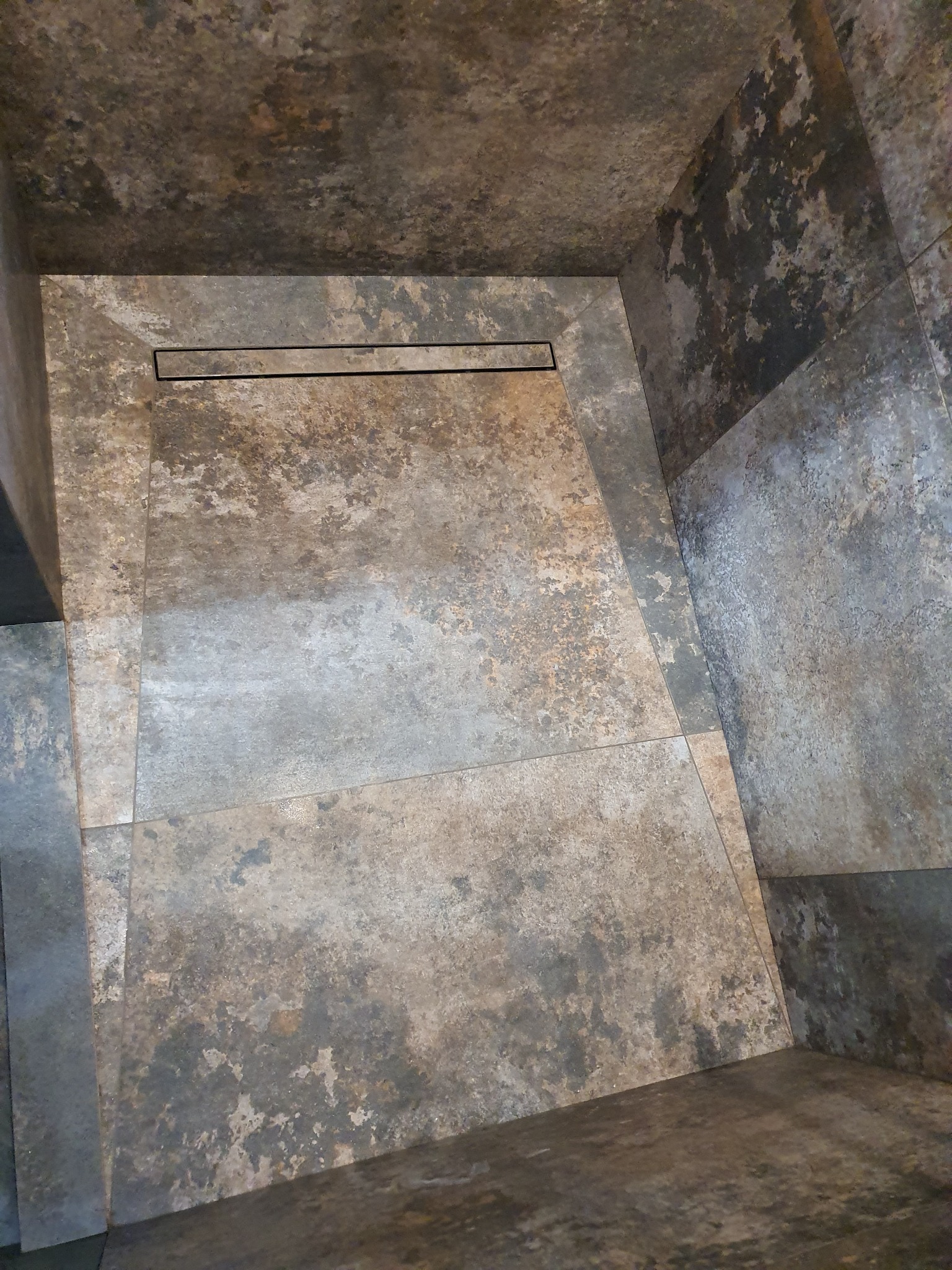 Nowoczesna kabina prysznicowa z odpływem liniowym, wykończona płytkami imitującymi beton w odcieniach szarości i brązu, widok z góry.