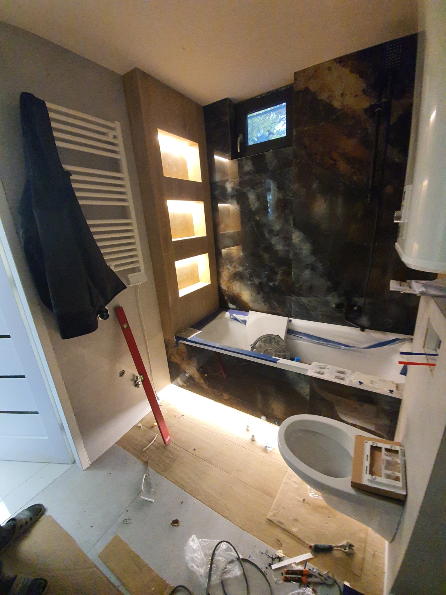 Remontowana łazienka z podświetlaną wnęką, czarną ścianą z efektem marmuru, białą wanną zabezpieczoną folią, oraz widocznym fragmentem toalety i narzędziami na podłodze w trakcie prac.