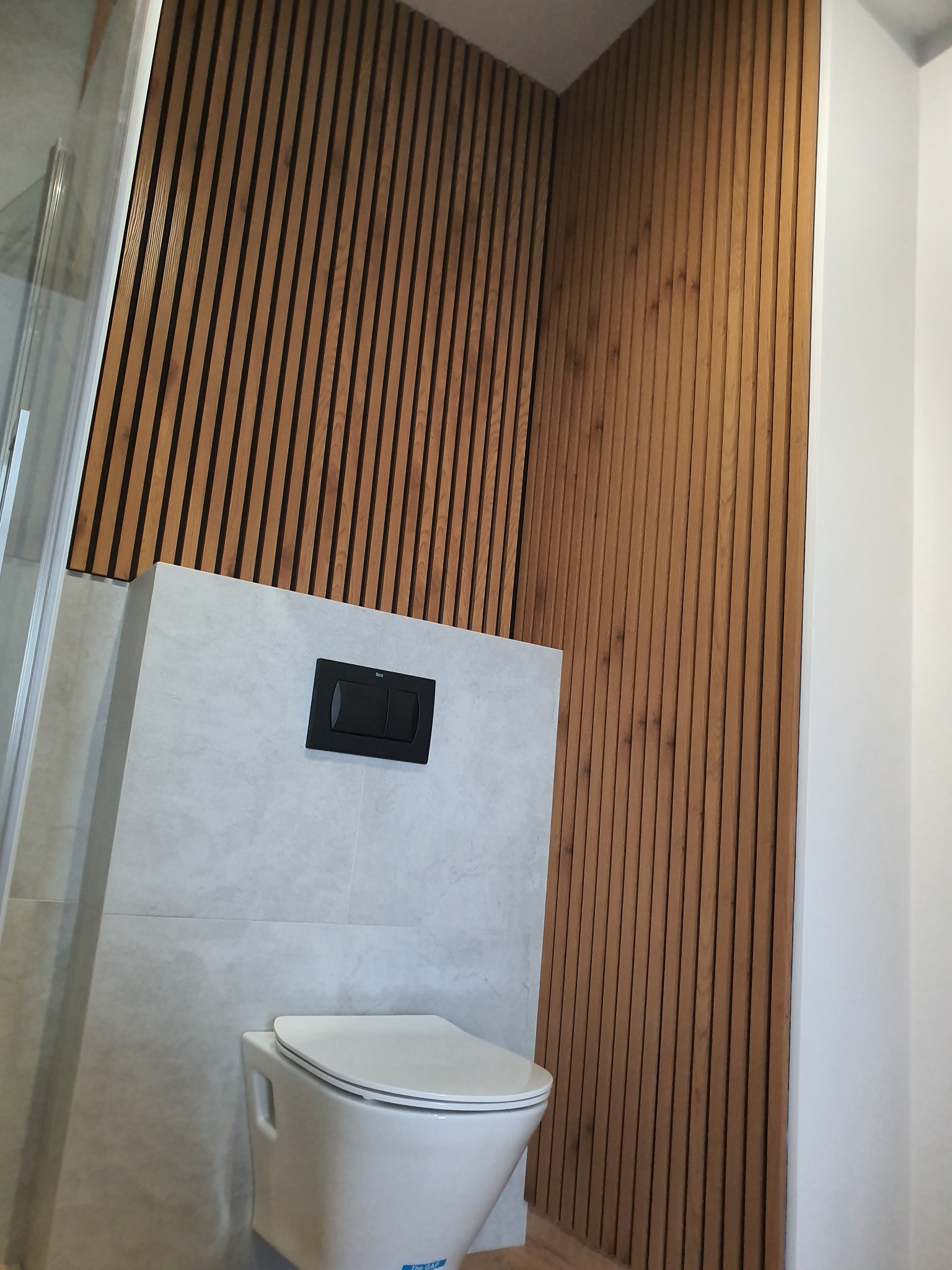 Nowoczesna łazienka z drewnianymi lamelami na ścianie, wisząca toaleta z czarnym przyciskiem spłukującym i szklana kabina prysznicowa w rogu.