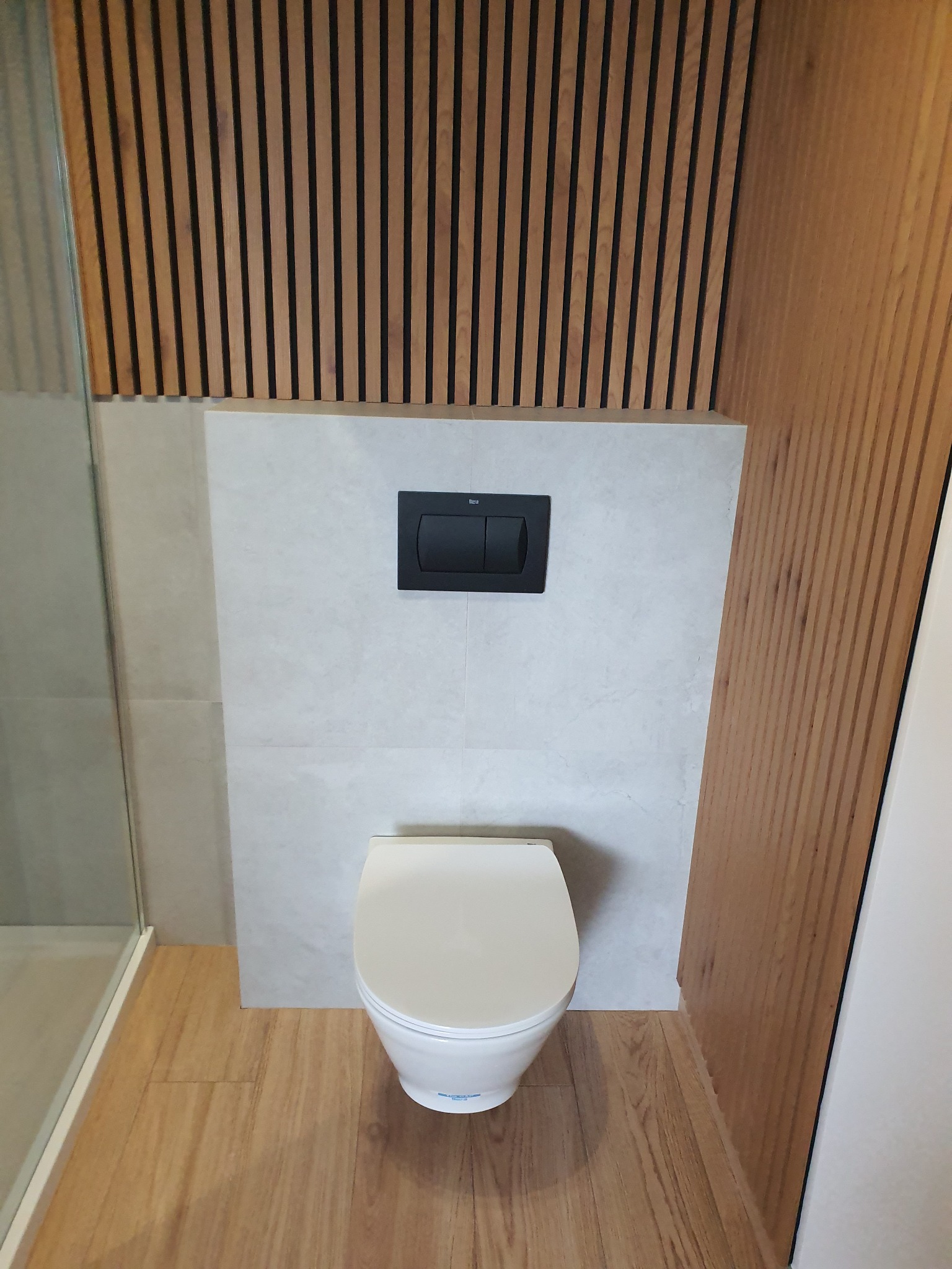 Nowoczesna toaleta podwieszana z czarnym przyciskiem spłukującym na tle szarej ściany z płytek, drewniana podłoga i dekoracyjne panele ścienne z drewna w łazience.
