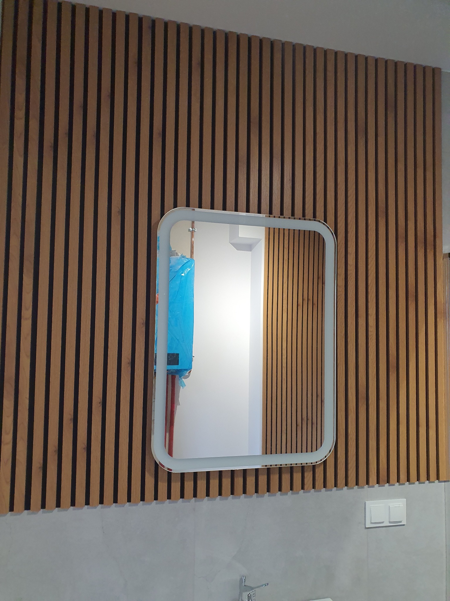 Nowoczesna łazienka z lustrem LED na ścianie z drewnianych lameli. W lustrze odbija się bojler w niebieskiej folii ochronnej.