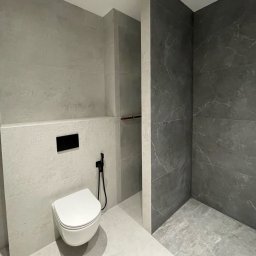 BudCapital - Nowoczesna łazienka z szarymi płytkami imitującymi beton i marmur, podwieszana toaleta z czarną armaturą bidetową, minimalistyczny design.