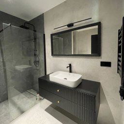 BudCapital - Nowoczesna łazienka z czarną armaturą, szklaną kabiną prysznicową i umywalką nablatową na szafce z frezowanymi frontami. Minimalistyczne oświetlenie nad lustrem.