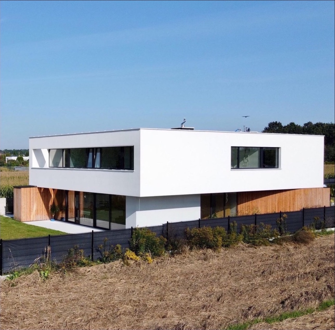 Nowoczesny, dwukondygnacyjny dom z białymi ścianami i drewnianą elewacją na parterze, otoczony zielenią i czarnym ogrodzeniem, w słoneczny dzień.