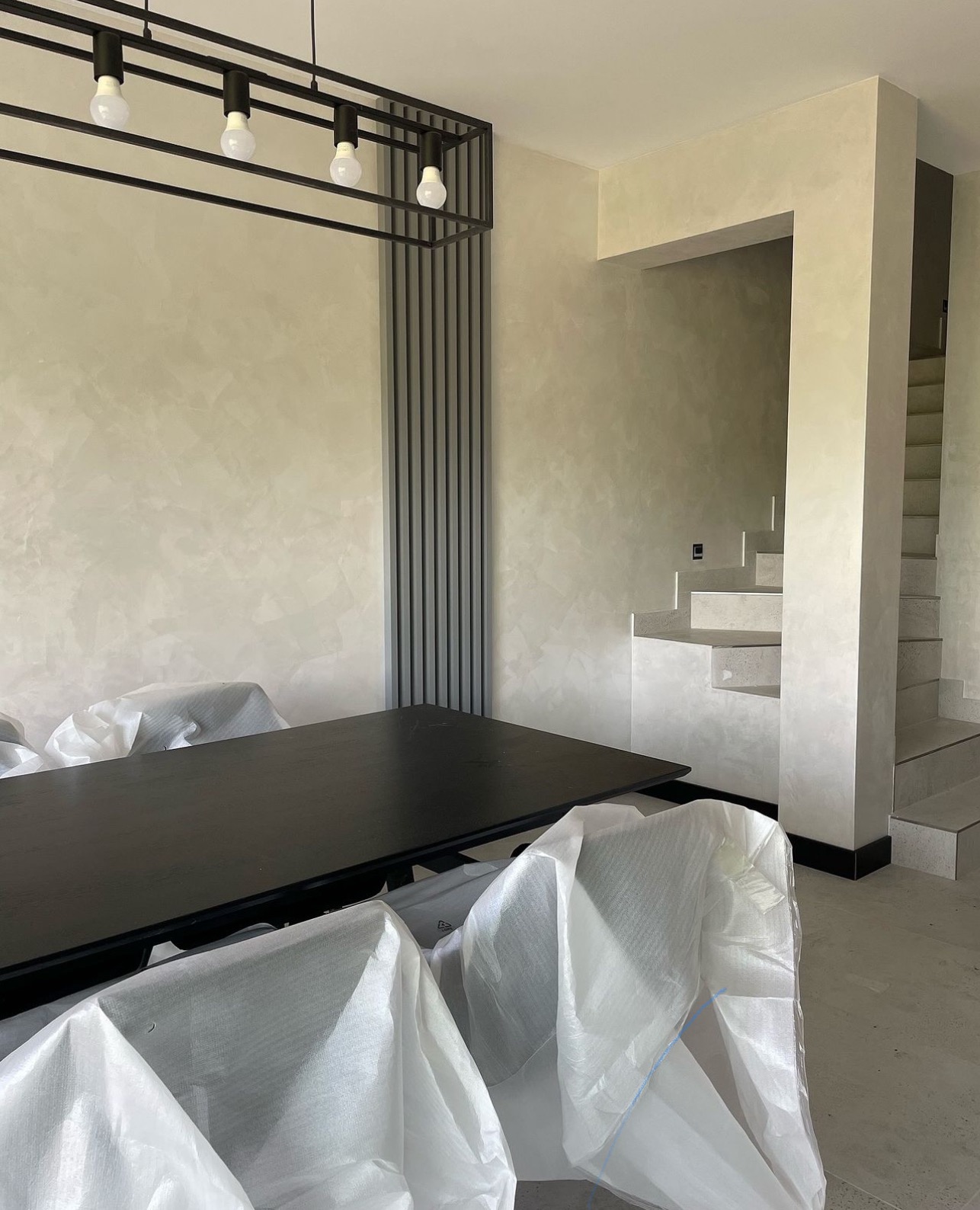 Wnętrze w Warszawie po malowaniu ścian z efektem dekoracyjnym, z widocznymi schodami i lampą sufitową, krzesła zabezpieczone folią ochronną.