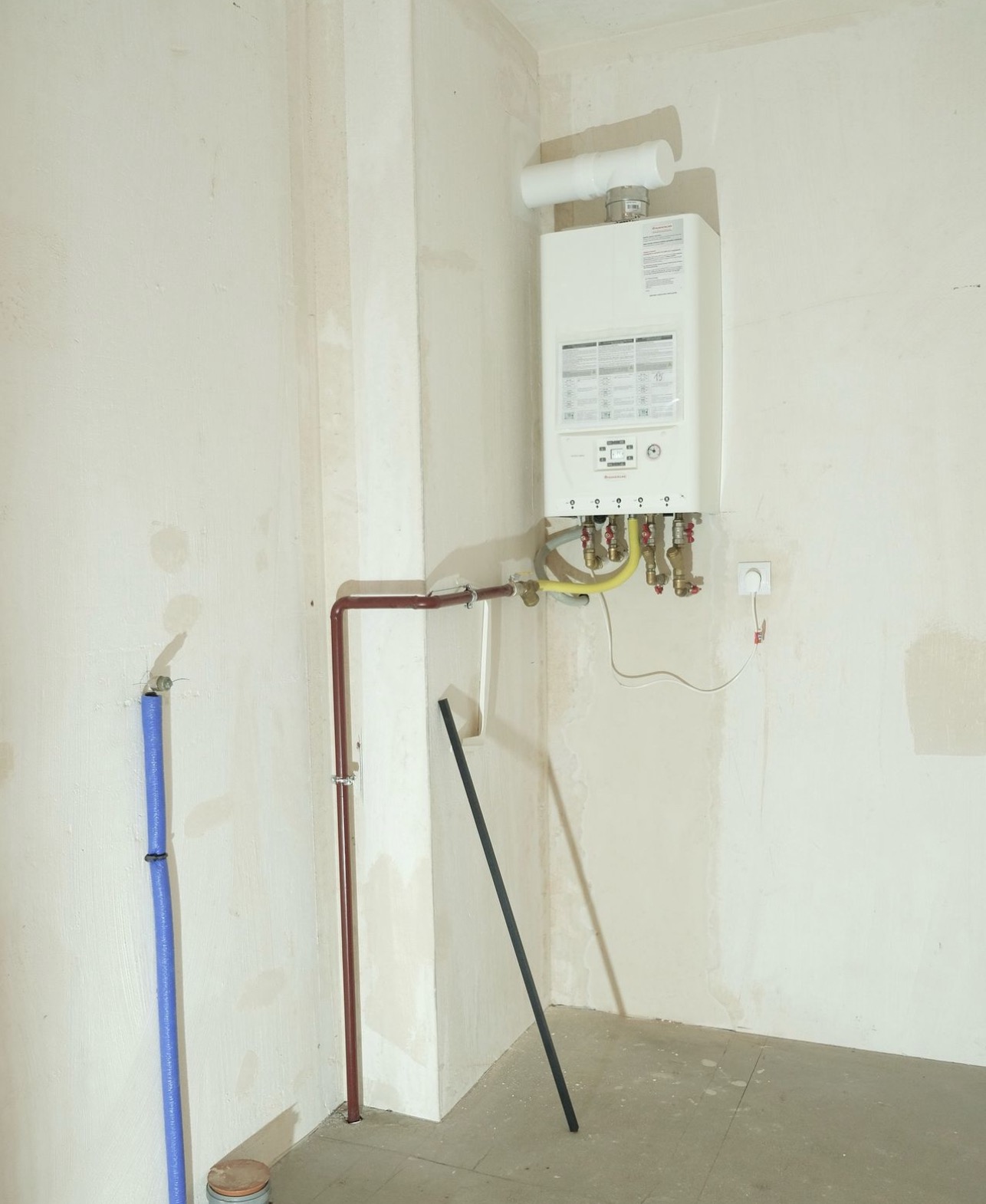 Biały piec gazowy kondensacyjny zamontowany na surowej ścianie z widocznymi instalacjami wodnymi i gazowymi, w tle nieotynkowany narożnik i podłoga w trakcie remontu.