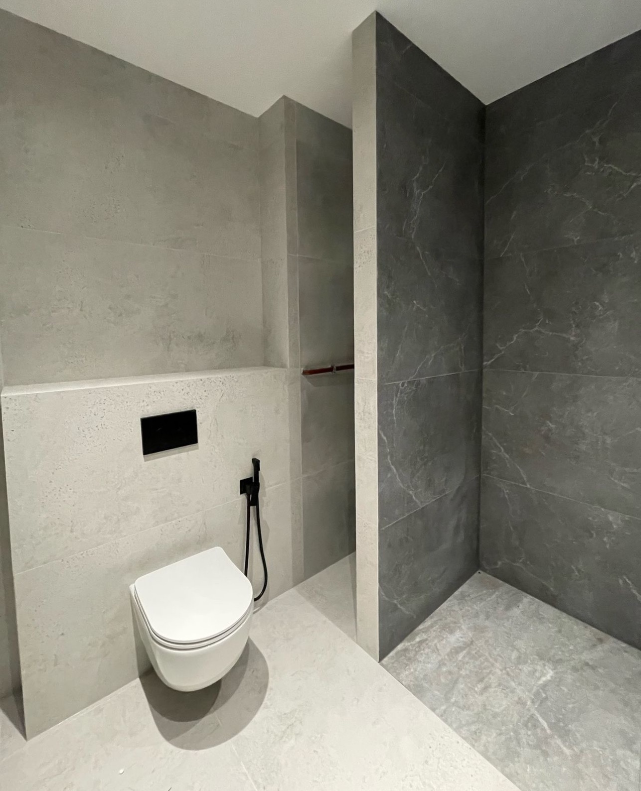 Nowoczesna łazienka z szarymi płytkami imitującymi beton i marmur, podwieszana toaleta z czarną armaturą bidetową, minimalistyczny design.