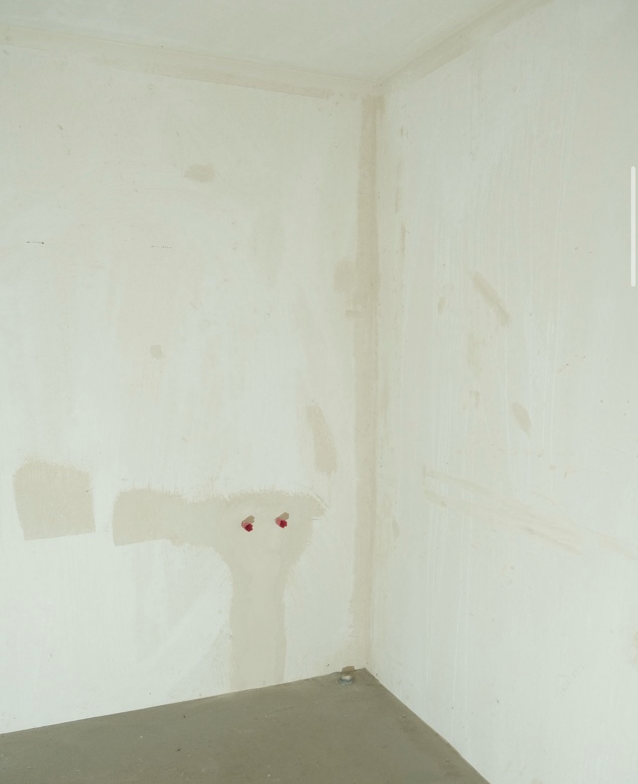 Narożnik pokoju w trakcie prac wykończeniowych: ściany po szpachlowaniu, widoczne ślady po instalacji elektrycznej zabezpieczone czerwonymi zaślepkami, przygotowane do dalszych prac.