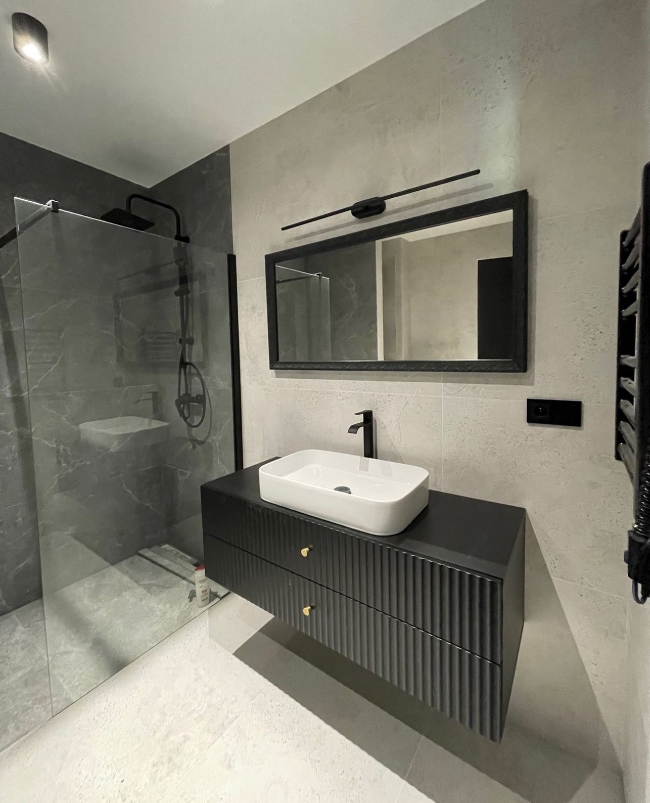 Nowoczesna łazienka z czarną armaturą, szklaną kabiną prysznicową i umywalką nablatową na szafce z frezowanymi frontami. Minimalistyczne oświetlenie nad lustrem.