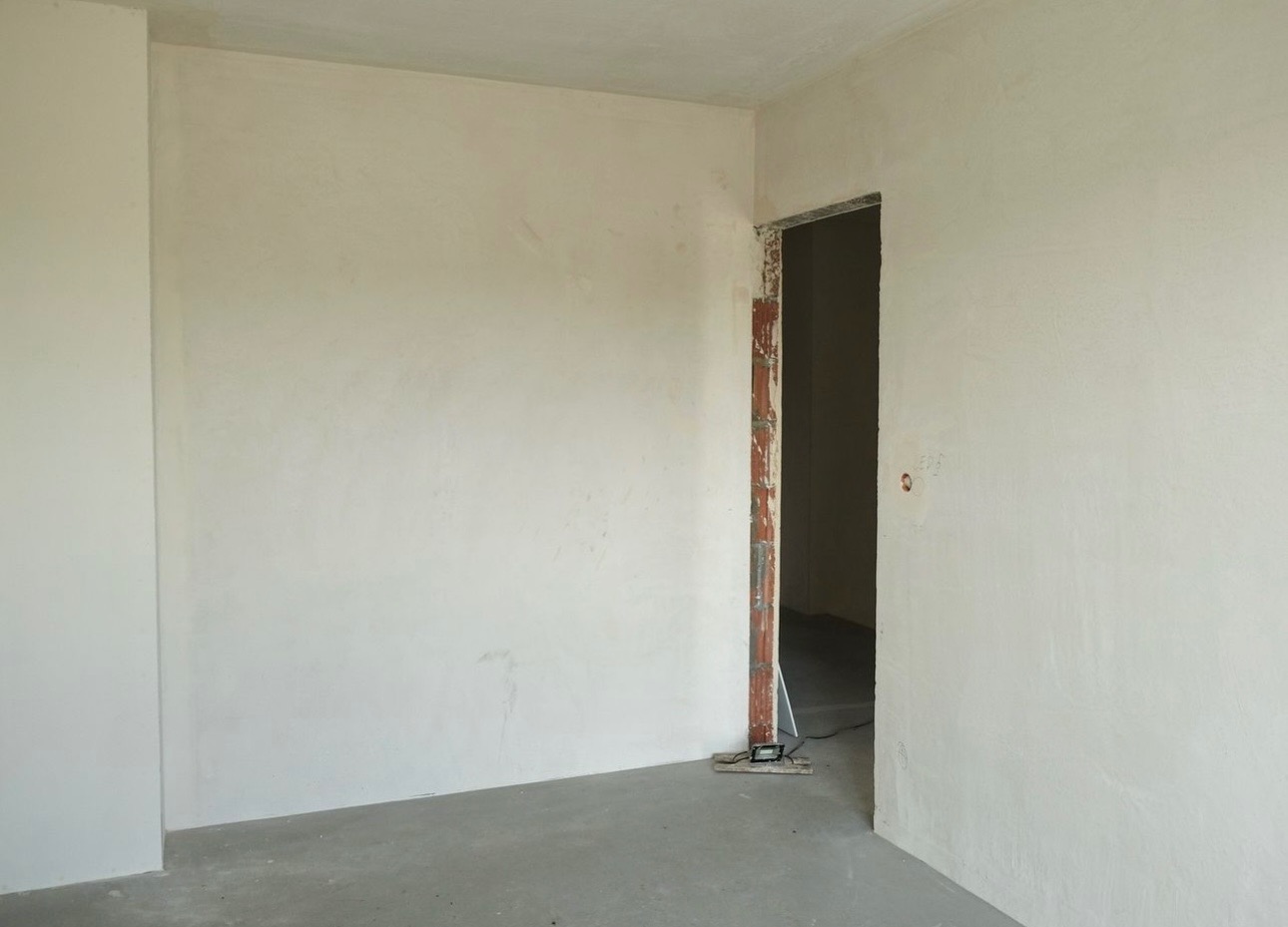 Surowe wnętrze w trakcie prac wykończeniowych: ściany wstępnie wygładzone gładzią, surowa ościeżnica drzwiowa, widoczne ślady po instalacjach elektrycznych.