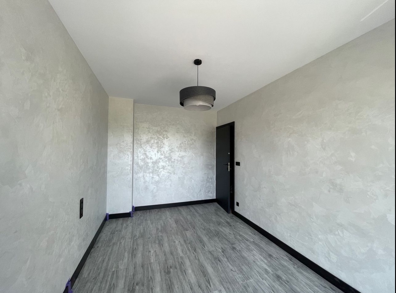 Wnętrze pokoju po malowaniu dekoracyjną farbą w odcieniach szarości, widoczne zabezpieczenia podłogi przy listwach, nowoczesna lampa sufitowa i czarne drzwi.
