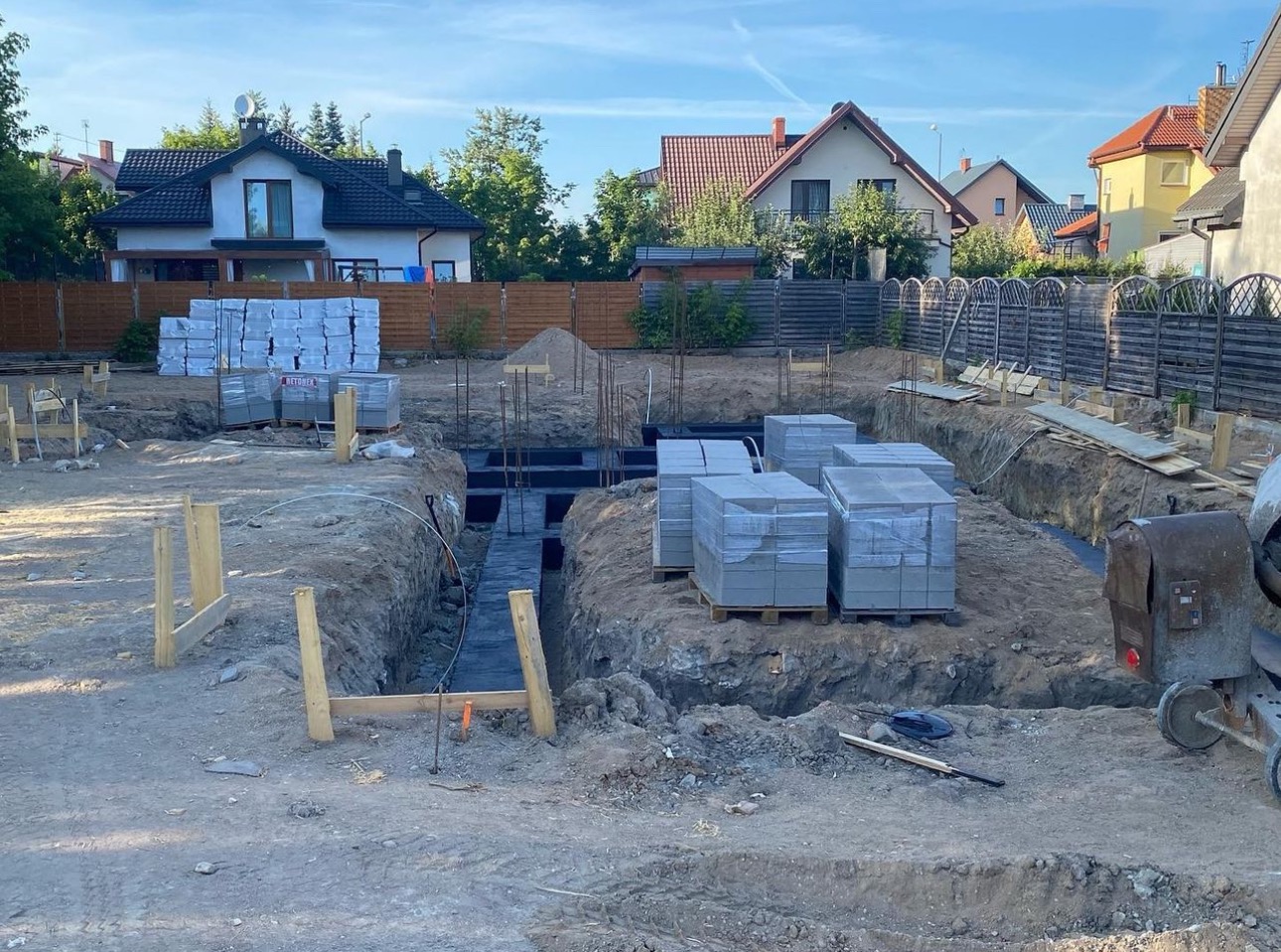 Fundamenty domu jednorodzinnego w trakcie budowy, widoczne wykopane rowy, zbrojenie i bloczki betonowe na paletach, w tle zabudowa jednorodzinna.