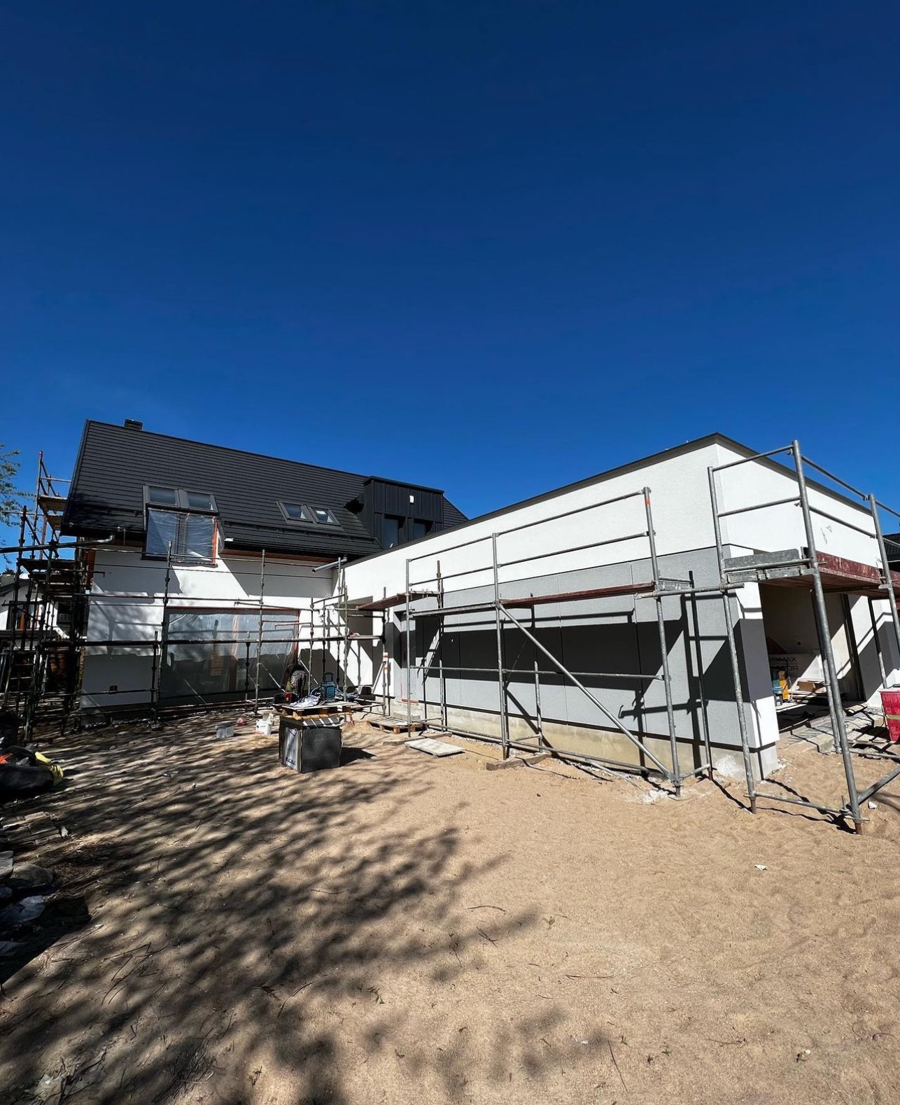 Rozbudowa domu jednorodzinnego z widocznymi rusztowaniami i częściowo otynkowaną elewacją w kolorach bieli i szarości, pod błękitnym niebem.