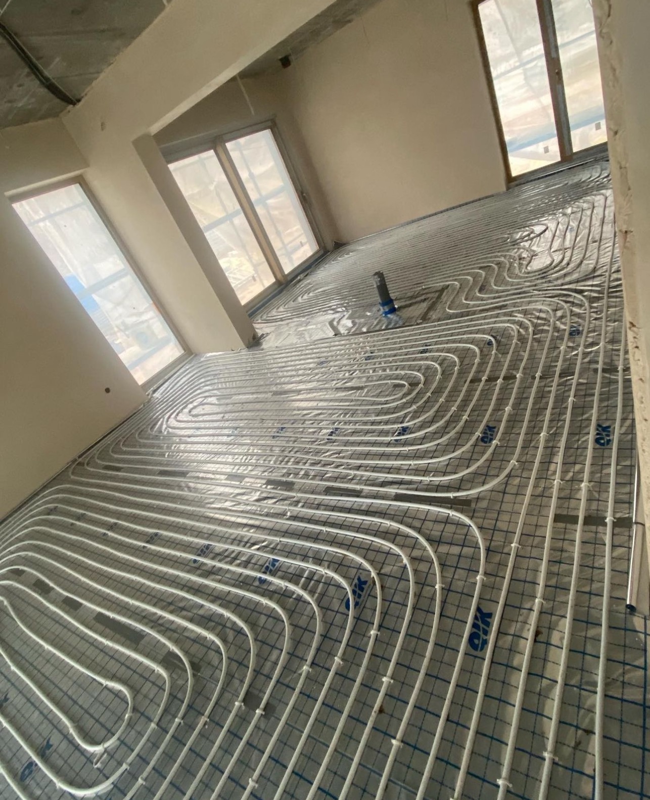 Rozłożone rury ogrzewania podłogowego na folii z siatką w jasnym, surowym wnętrzu z oknami.