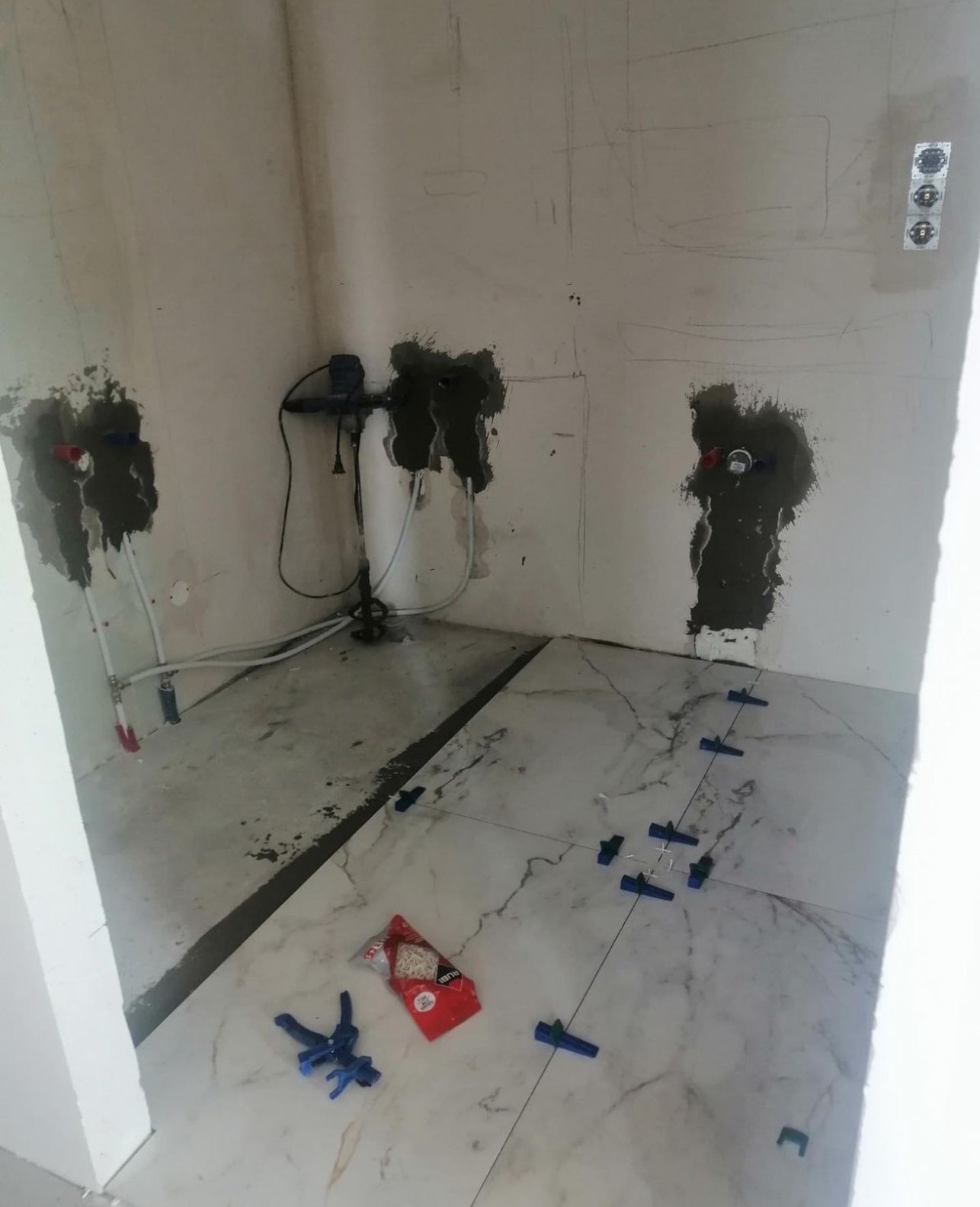 Wnętrze łazienki w trakcie remontu: ułożone płytki imitujące marmur, widoczne krzyżyki glazurnicze, przygotowane punkty instalacji wodnej w ścianie, ślady po zaprawie, worek z klinami do płytek.