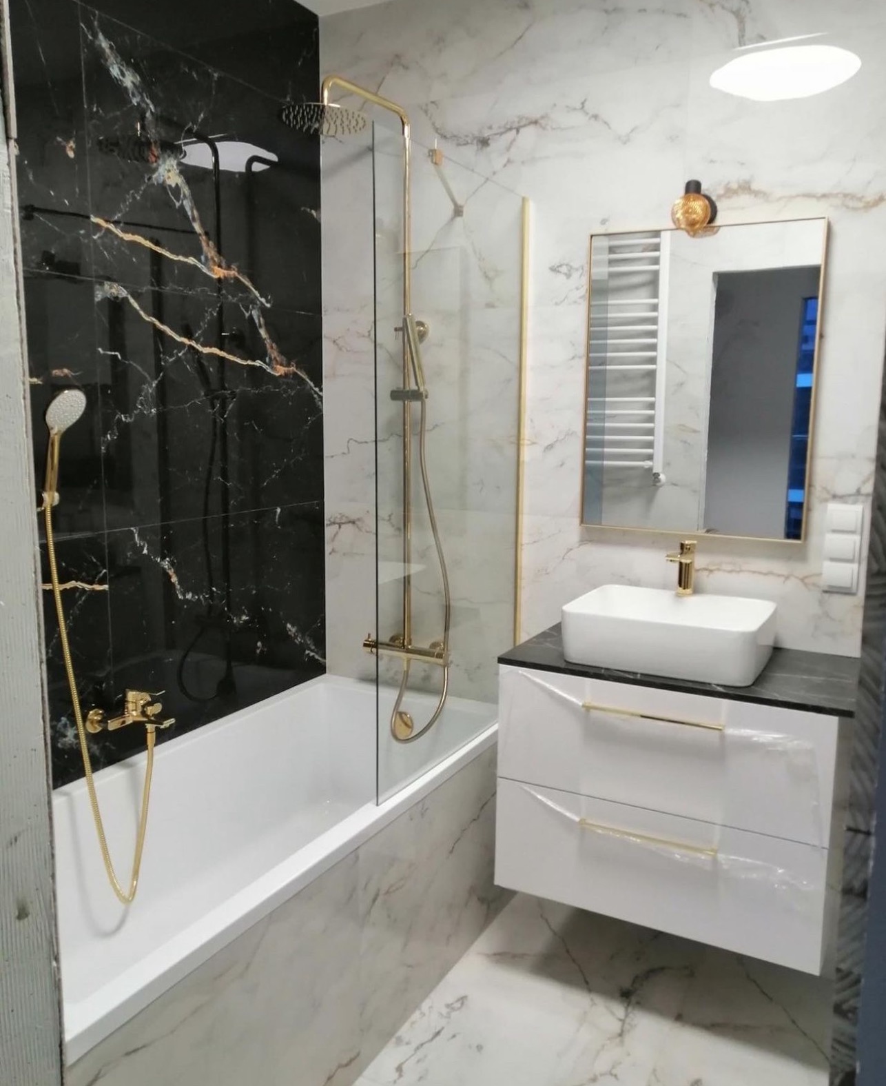 Nowoczesna łazienka z czarno-białymi marmurkowymi płytkami, złotą armaturą i białą szafką pod umywalką z czarnym blatem.