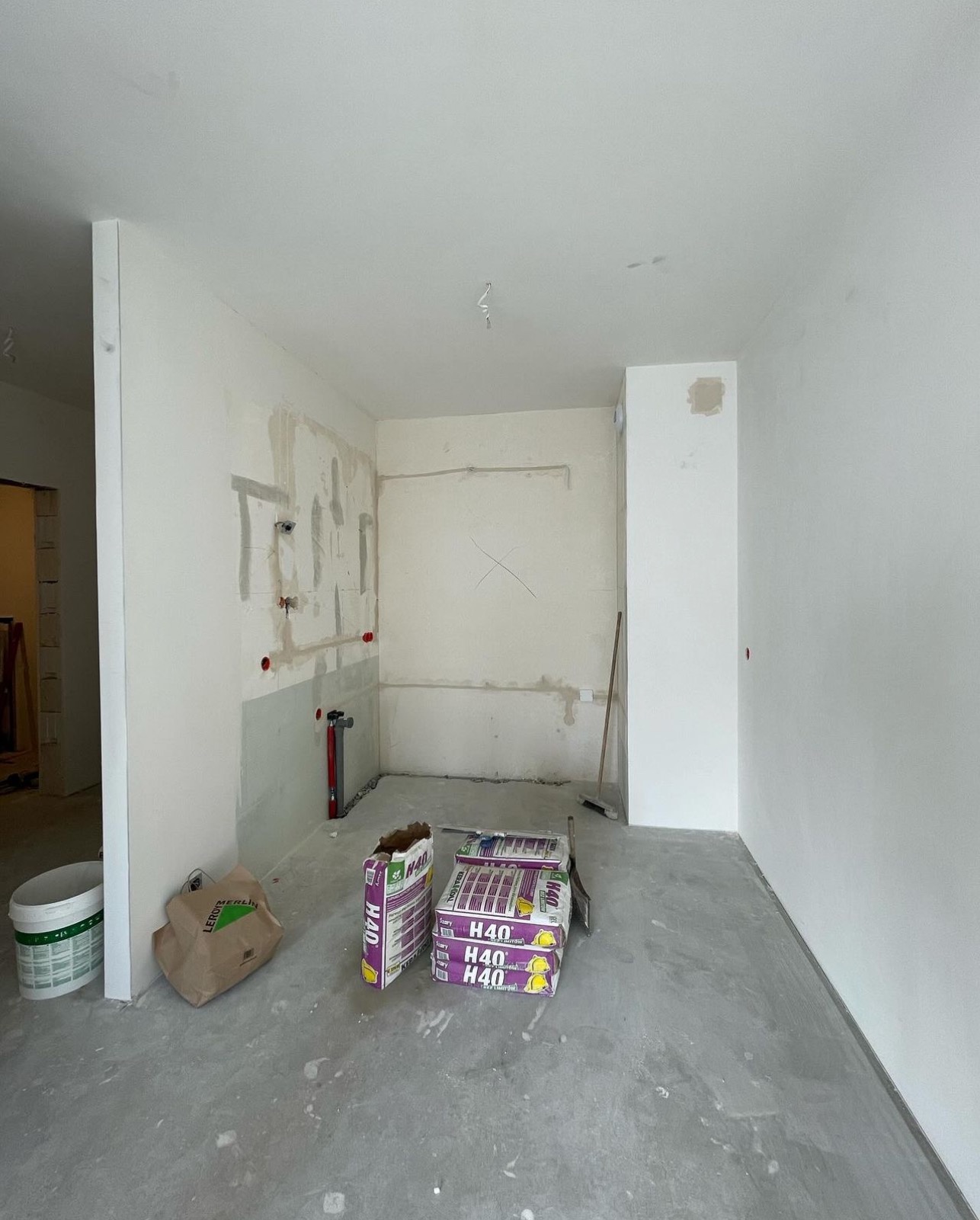 Wnętrze w trakcie prac wykończeniowych, widoczne przygotowane ściany, instalacje, worki z zaprawą klejową H40 oraz wiadro z farbą, podłoga w surowym betonie.