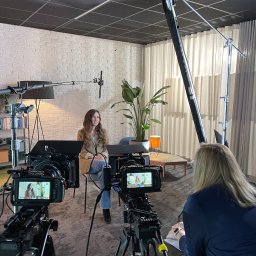 KO.REC MEDIA SPÓŁKA Z OGRANICZONĄ ODPOWIEDZIALNOŚCIĄ - Kobieta siedząca na krześle podczas nagrywania, widoczna przez dwa ekrany kamer, oświetlenie studyjne, biała ceglana ściana w tle.