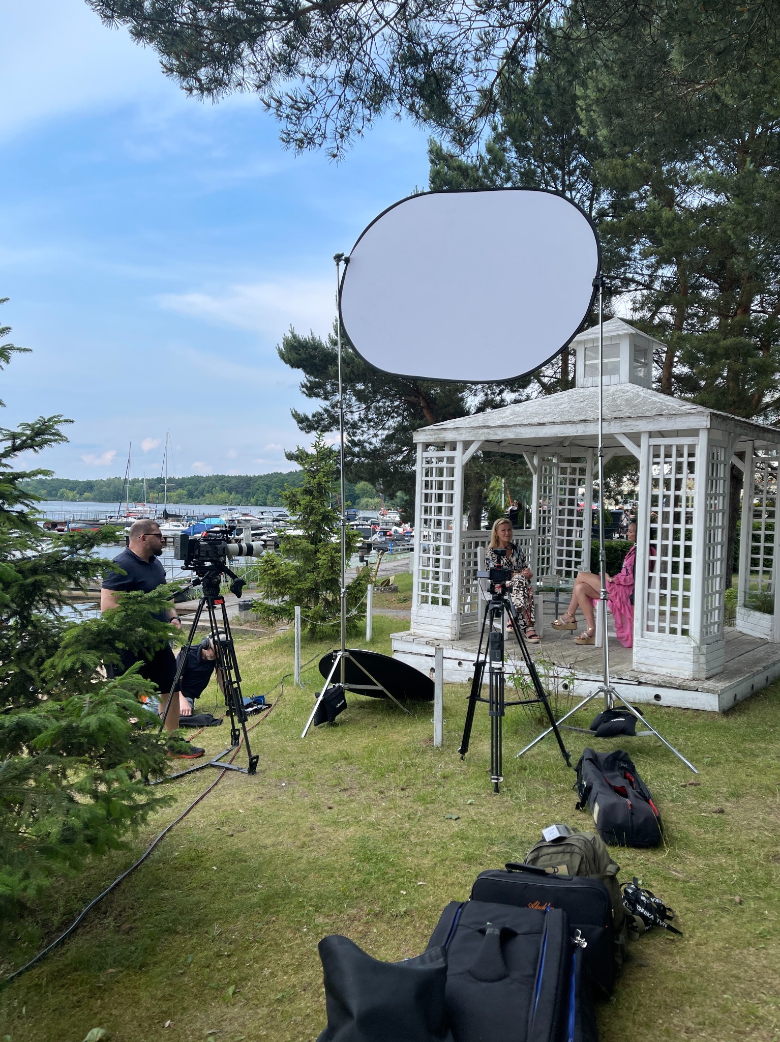 Plenerowa realizacja podcastu: dwie kobiety siedzą w białej altanie, nagrywane przez operatora z użyciem trzech kamer i blendy, w tle jezioro z żaglówkami.