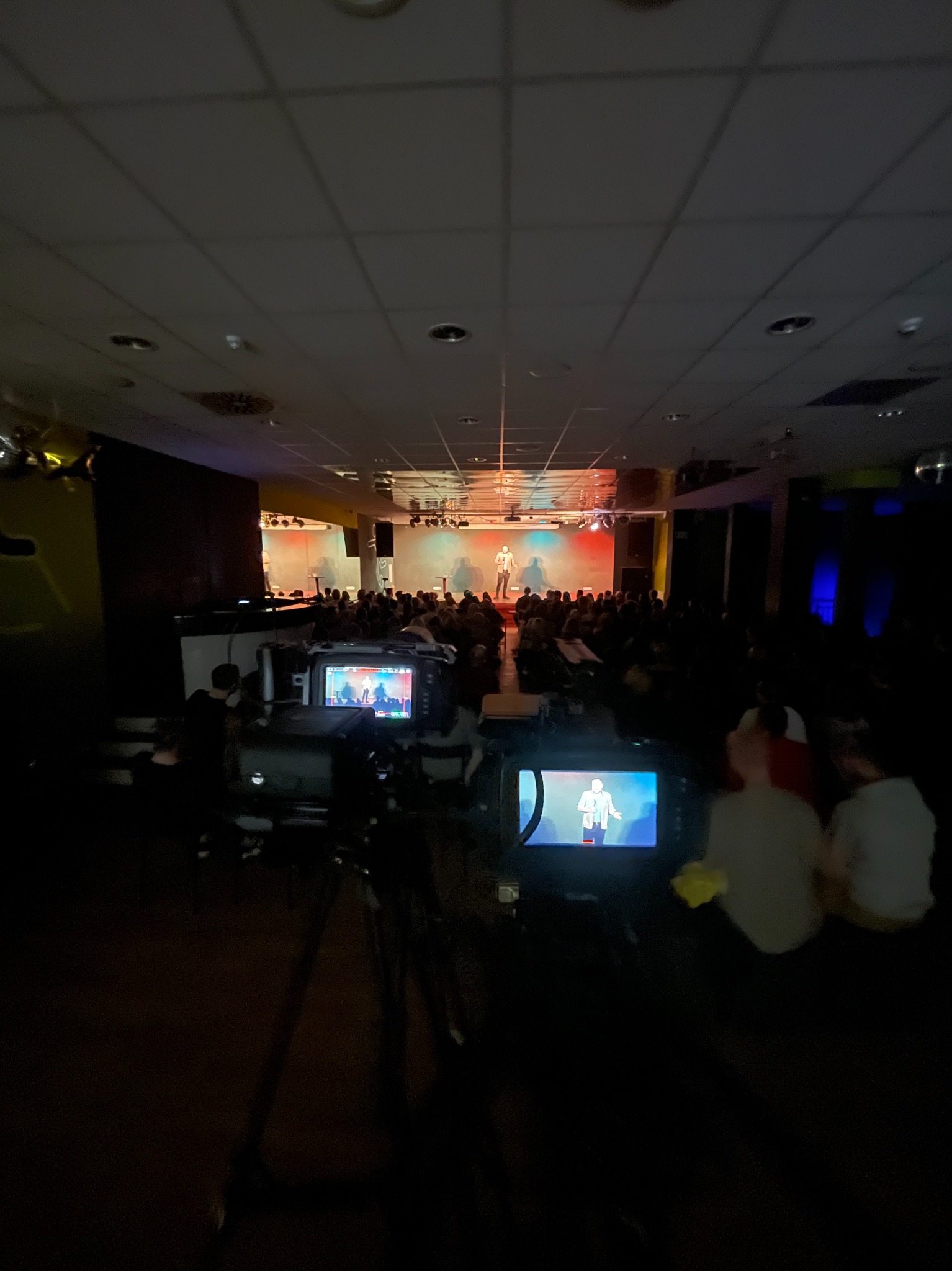 Kamera ustawiona na statywie rejestruje występ na scenie przed publicznością; widoczny ekran kamery z podglądem obrazu.