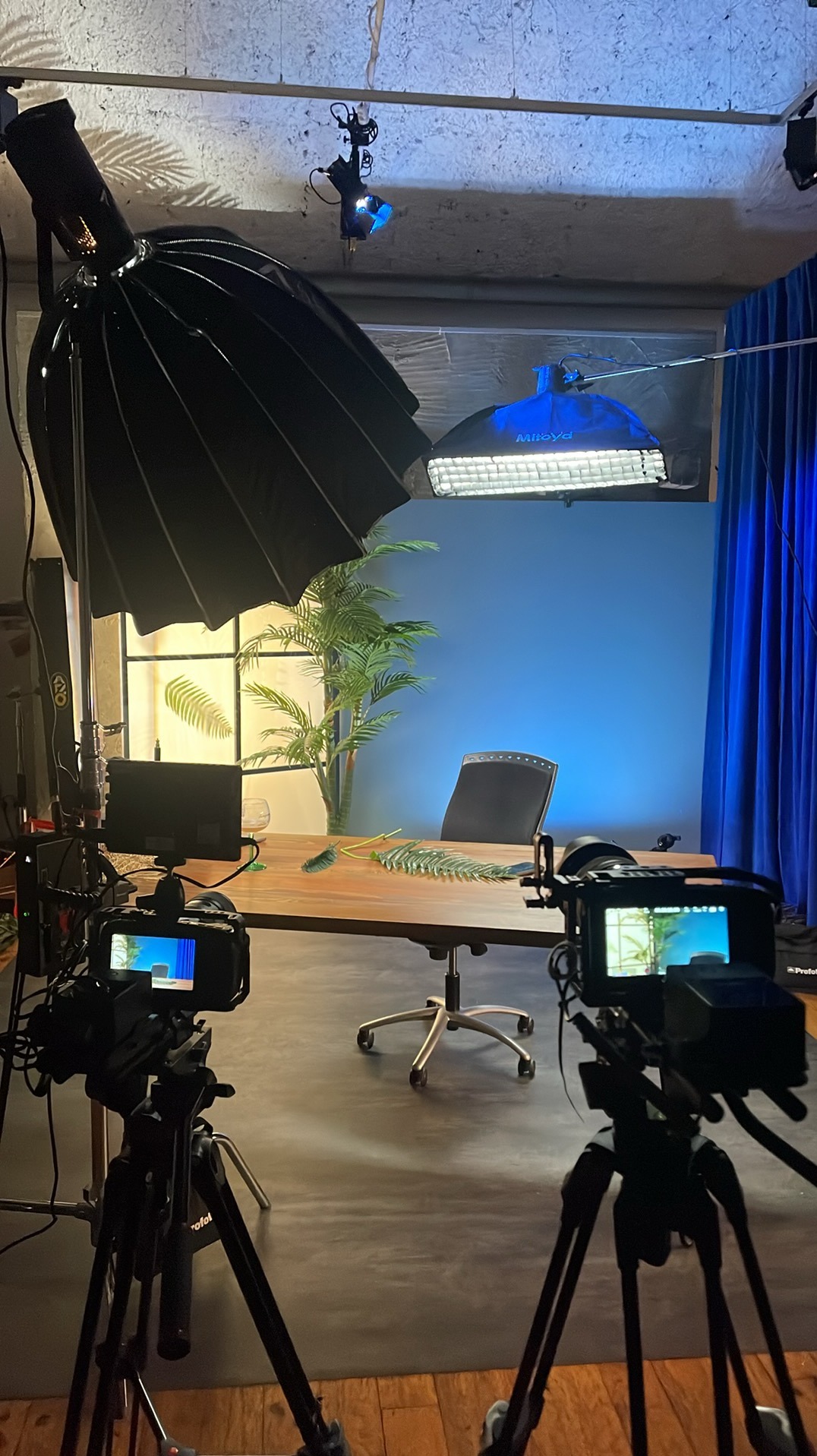 Dwa aparaty na statywach ustawione w studio filmowym z oświetleniem, biurkiem, krzesłem i dekoracjami, przygotowane do nagrywania.