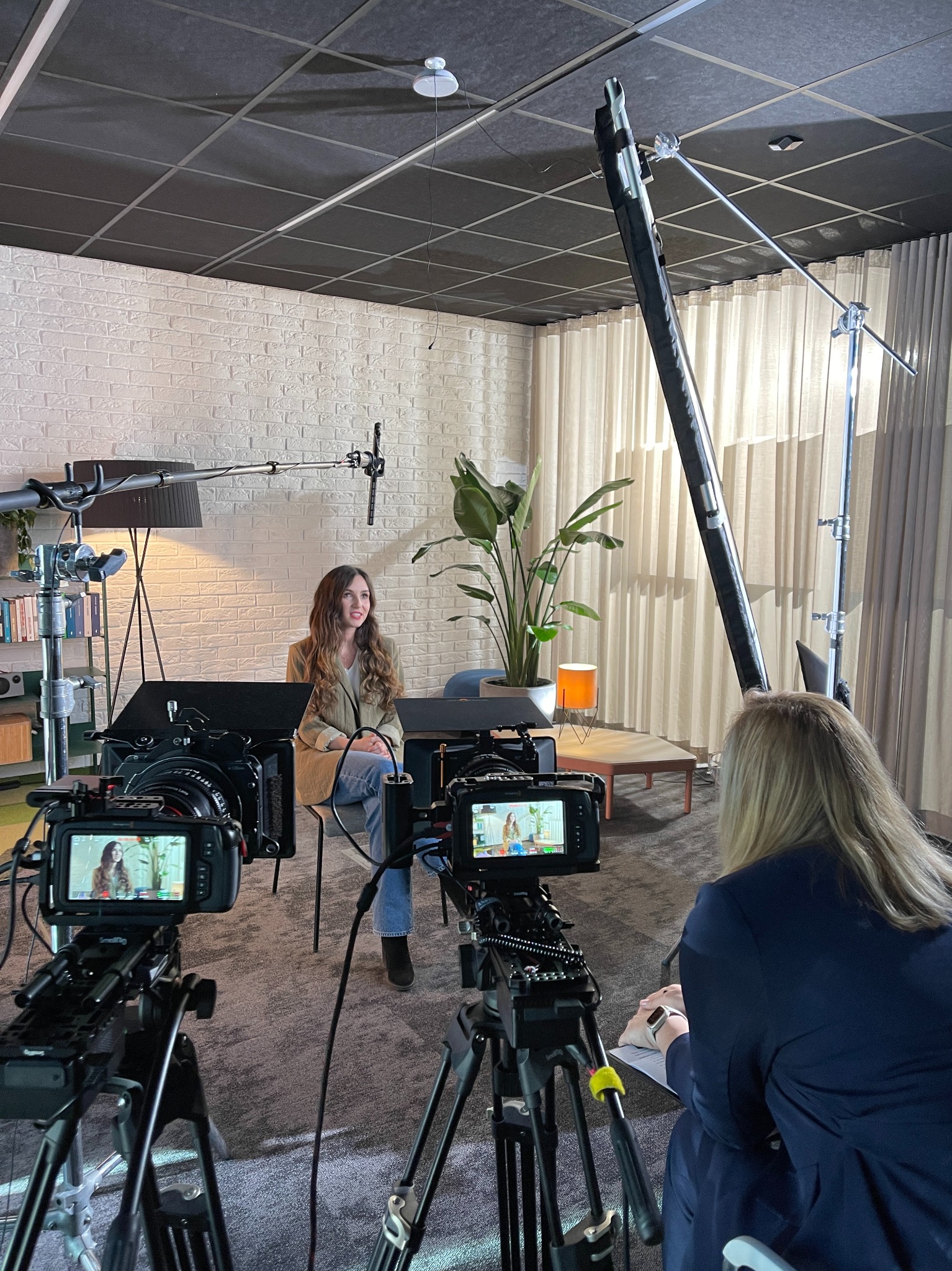 Kobieta siedząca na krześle podczas nagrywania, widoczna przez dwa ekrany kamer, oświetlenie studyjne, biała ceglana ściana w tle.