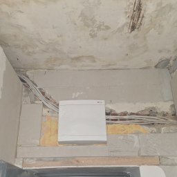 DUDI-ELEKTRO ŁUKASZ DUDEK - Biała skrzynka elektryczna ETI w folii ochronnej, zamontowana na ścianie w trakcie remontu. Widoczne kable, surowy tynk i notatki na ścianie.
