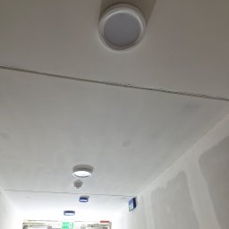 DUDI-ELEKTRO ŁUKASZ DUDEK - Biały sufit korytarza z zamontowanymi okrągłymi lampami sufitowymi LED i oznaczeniami ewakuacyjnymi. Widoczne ślady po szpachlowaniu.