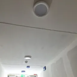 Biały sufit korytarza z zamontowanymi okrągłymi lampami sufitowymi LED i oznaczeniami ewakuacyjnymi. Widoczne ślady po szpachlowaniu.