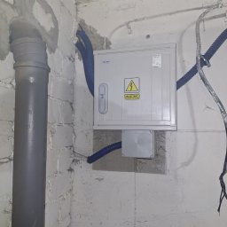 DUDI-ELEKTRO ŁUKASZ DUDEK - Szara skrzynka elektryczna PELMET z żółtym znakiem ostrzegawczym na ścianie z instalacją elektryczną w nieotynkowanym pomieszczeniu.