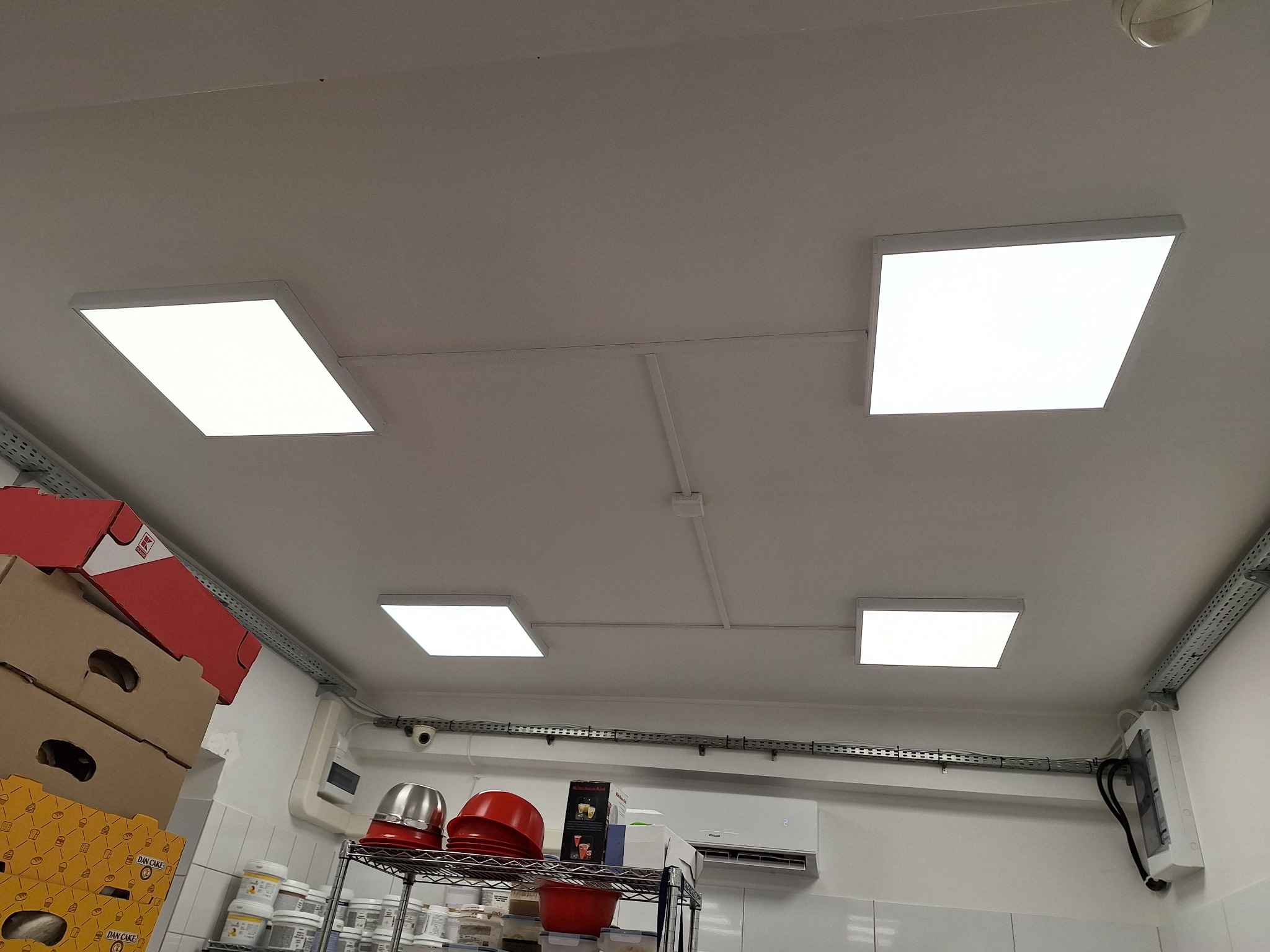 Cztery kwadratowe panele LED zamontowane na suficie w pomieszczeniu z elementami wyposażenia kuchennego, widoczne rury instalacyjne i klimatyzacja