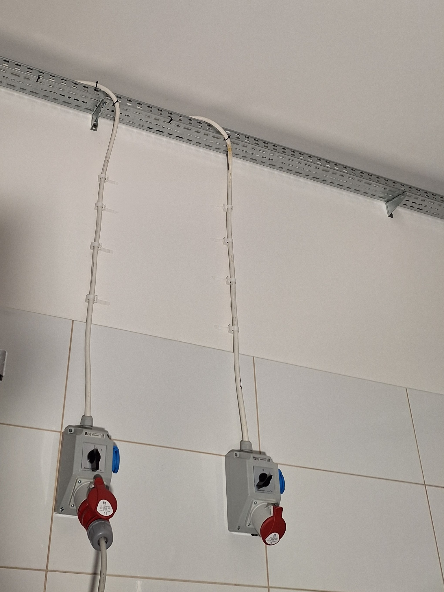 Dwa gniazda przemysłowe z wyłącznikami zamontowane na kafelkowanej ścianie, kable przymocowane do metalowej listwy kablowej pod sufitem za pomocą opasek zaciskowych.