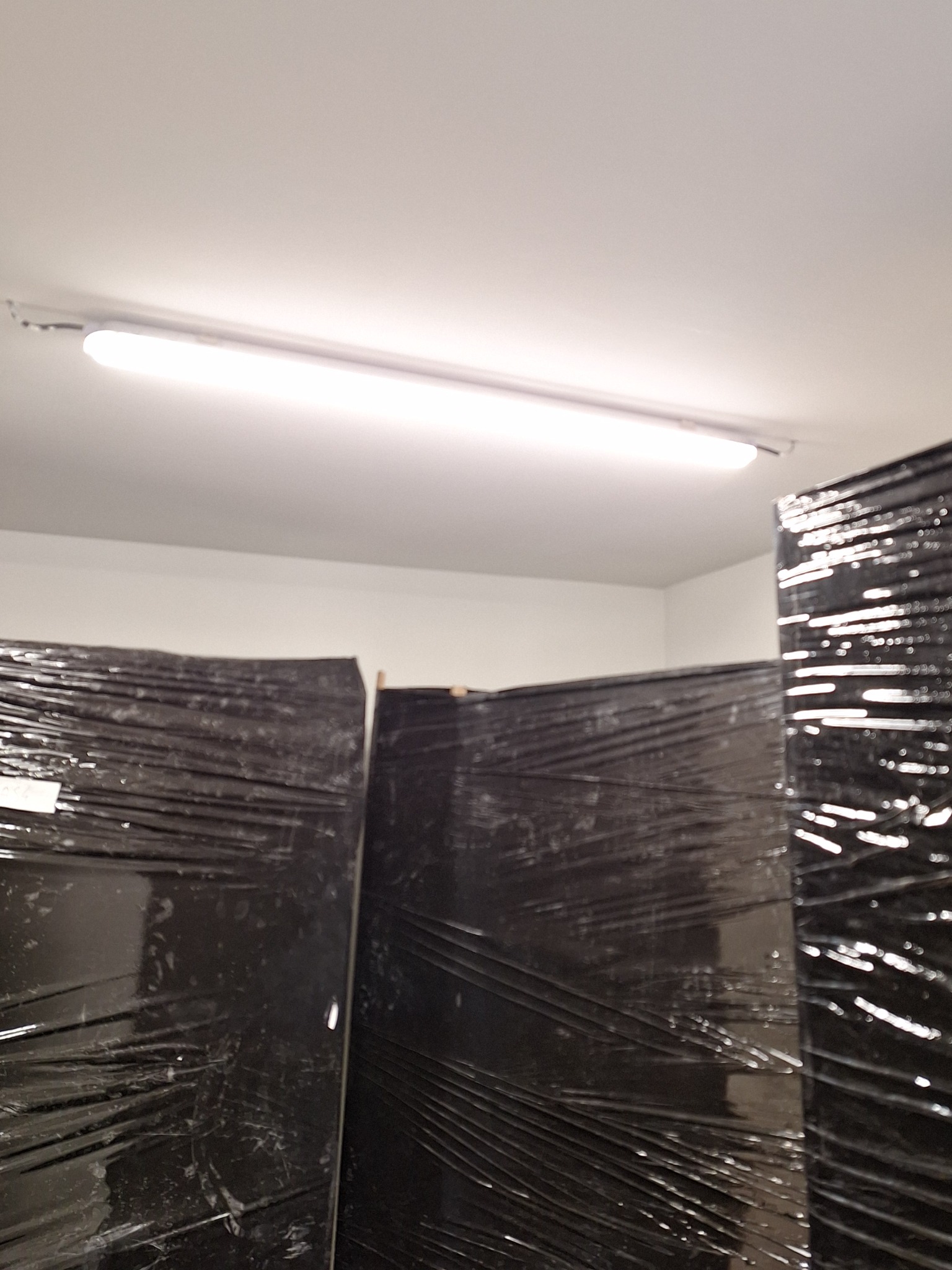 Paczki mebli owinięte czarną folią stretch, składowane w magazynie pod jarzeniówką.