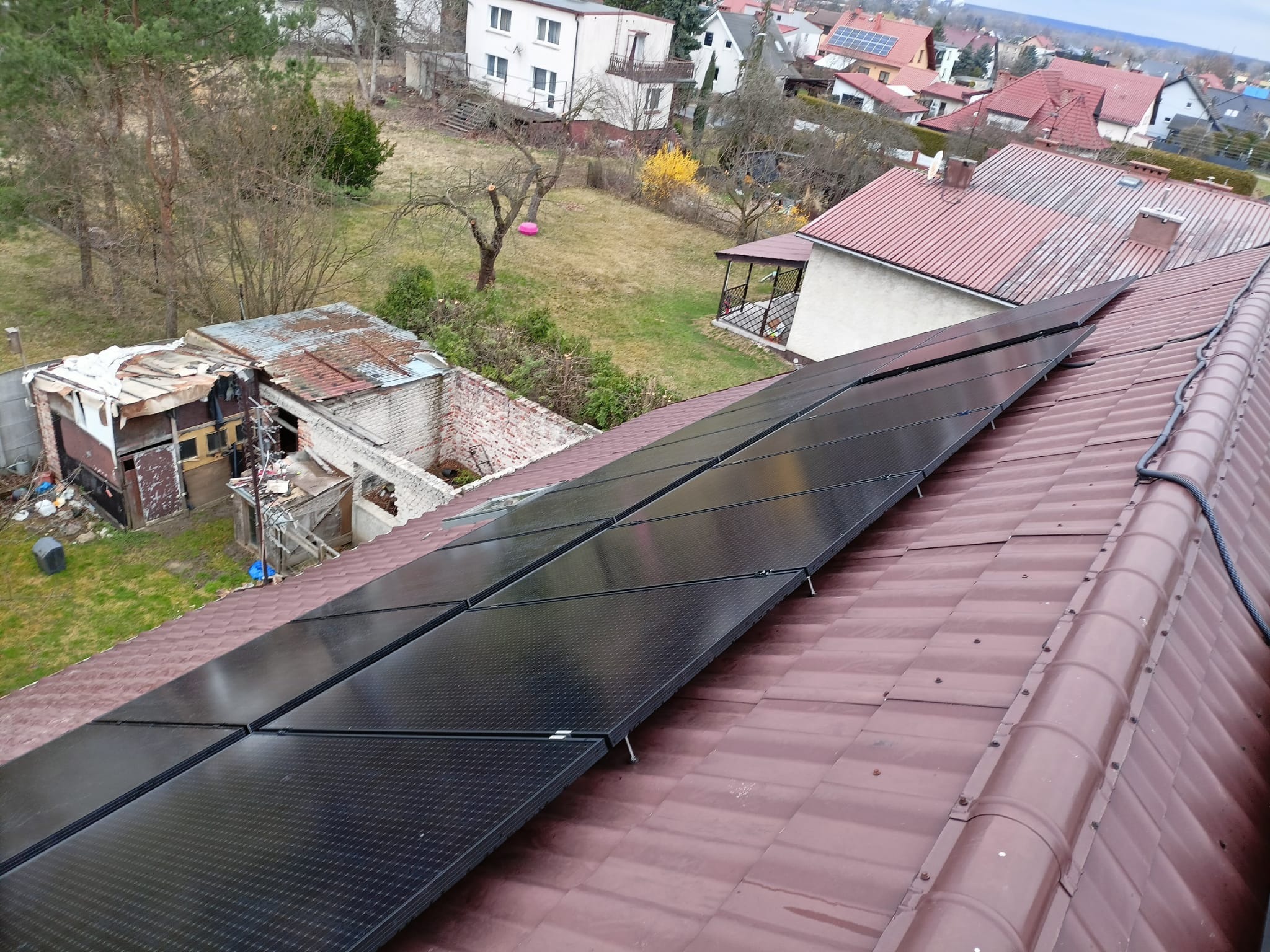 Panele fotowoltaiczne zamontowane na brązowym dachu z dachówki, widok z góry na okoliczne domy i ogród.