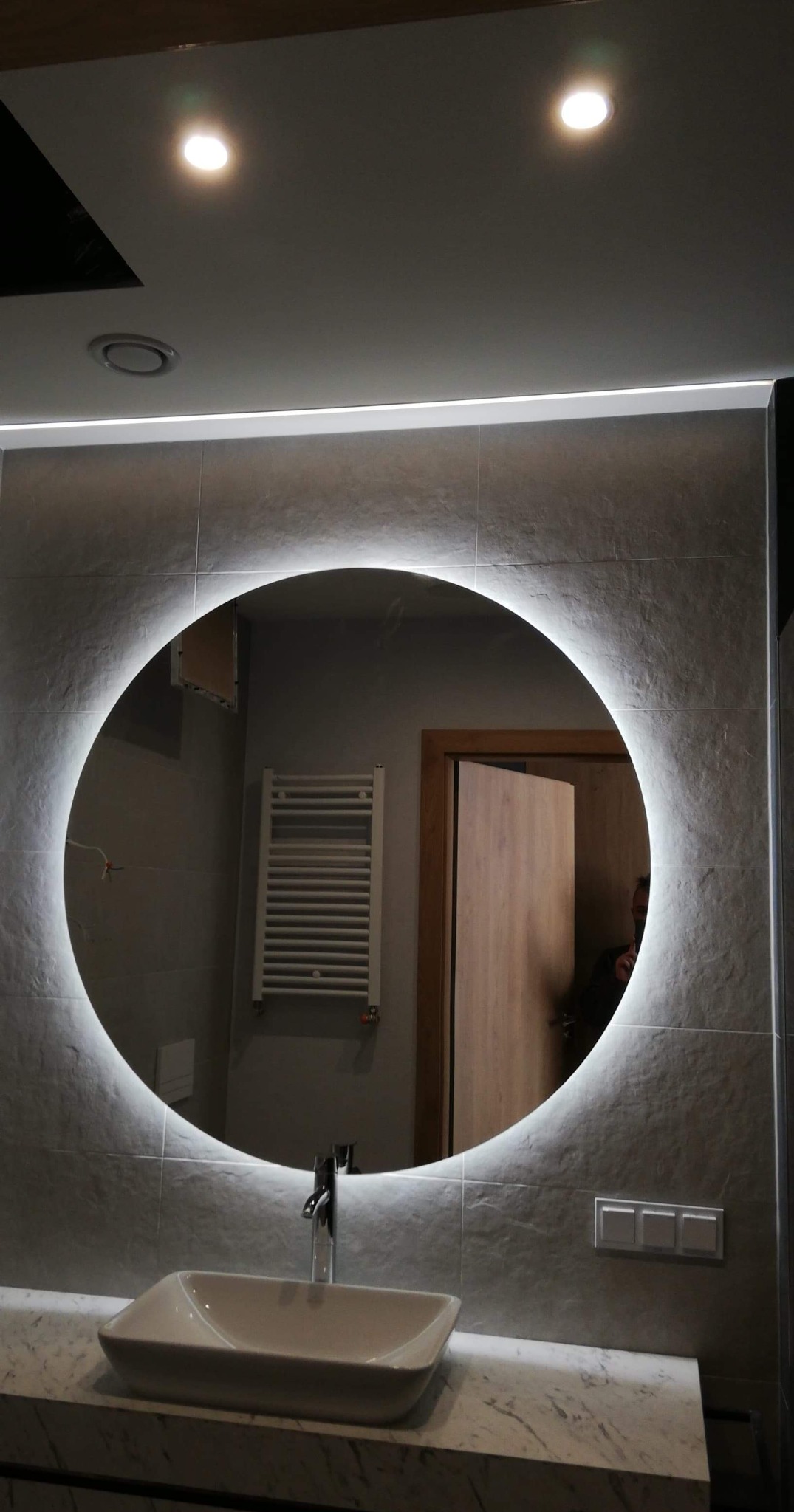 Nowoczesna łazienka z okrągłym lustrem podświetlanym LED, umywalką nablatową i szarą płytką imitującą kamień na ścianie.