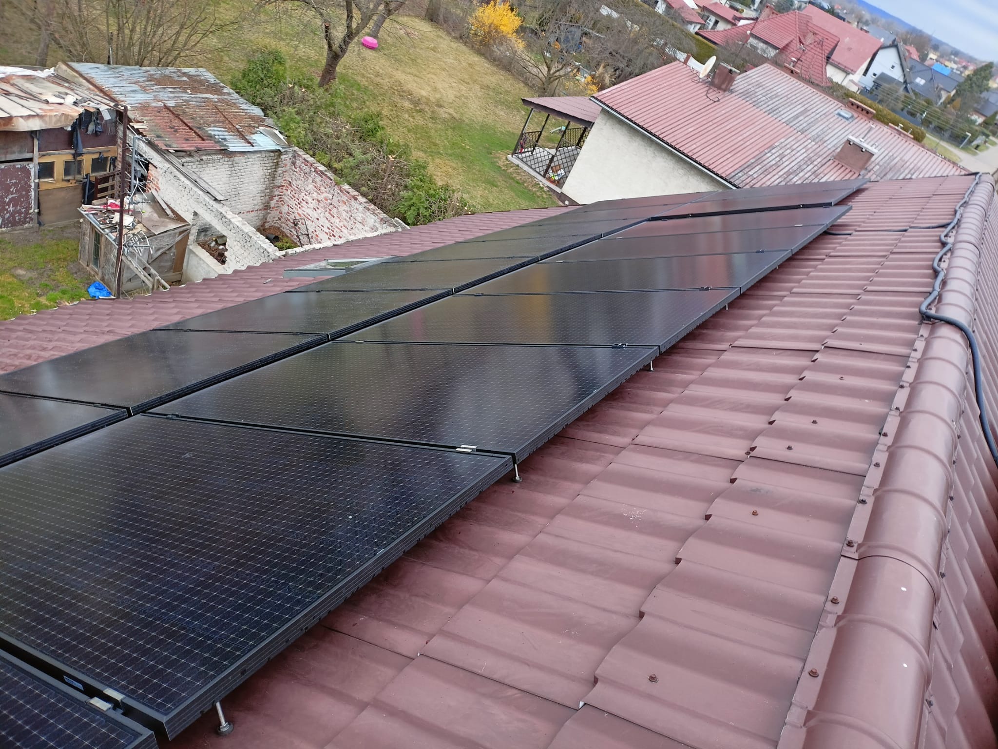 Rząd paneli słonecznych zamontowanych na brązowym dachu krytym dachówką ceramiczną, widok z góry na tle zabudowań mieszkalnych i zieleni.