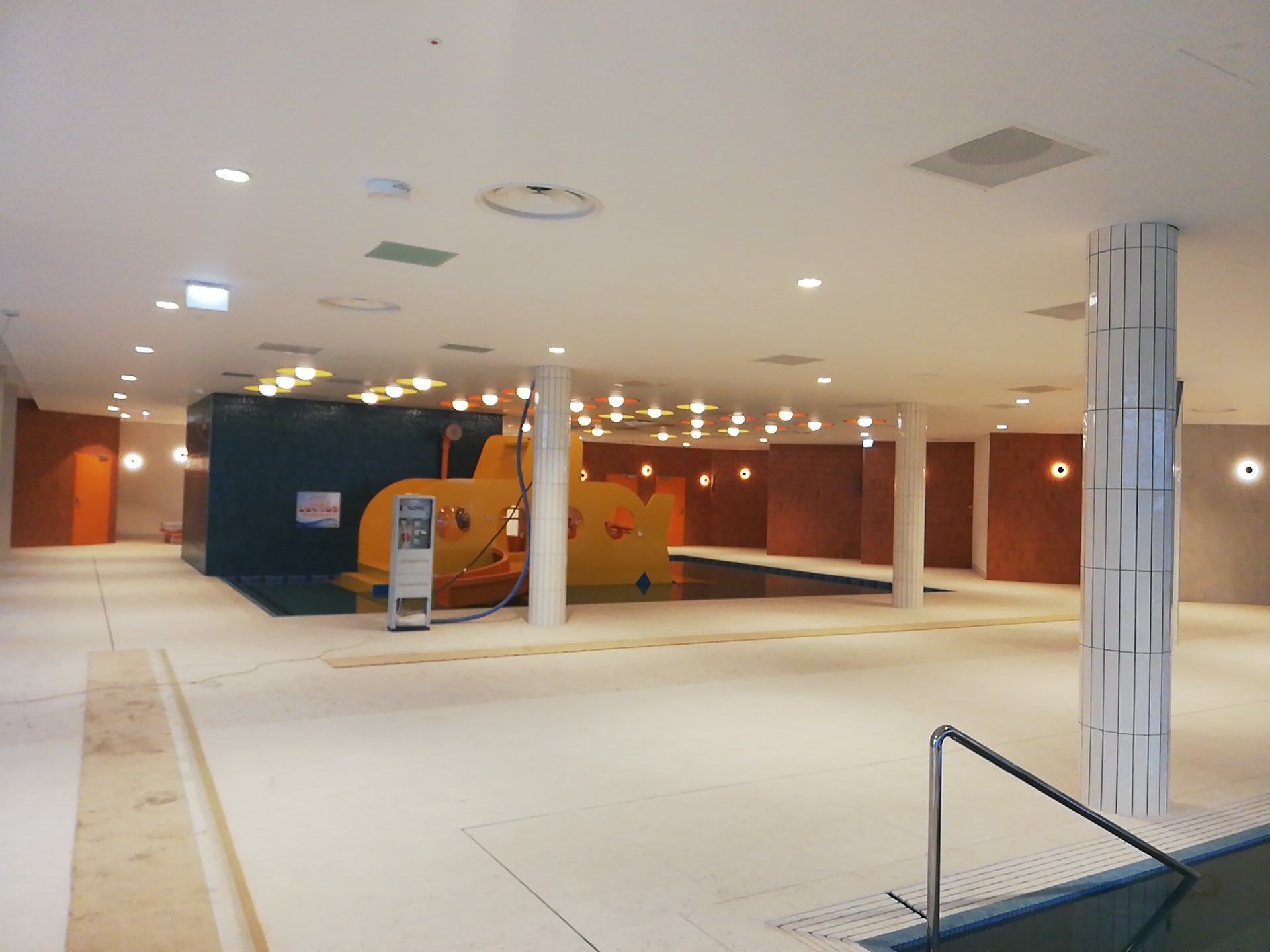 Wnętrze pustego basenu z żółtą konstrukcją w kształcie łodzi podwodnej, oświetleniem punktowym i kolumnami wyłożonymi białymi płytkami.