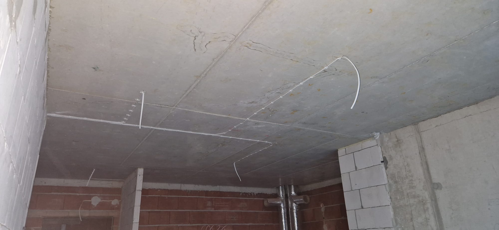 Surowy betonowy sufit z widocznymi przewodami elektrycznymi poprowadzonymi w korytkach i bezpośrednio po powierzchni, ściany z cegły i bloczków betonowych w stanie deweloperskim.