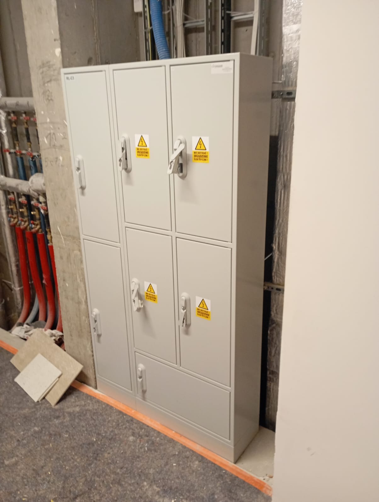 Szara szafa elektryczna z żółtymi ostrzeżeniami przed porażeniem prądem, umieszczona w surowym wnętrzu budynku w trakcie wykończenia, widoczne rury instalacji grzewczej i wentylacyjnej w tle.
