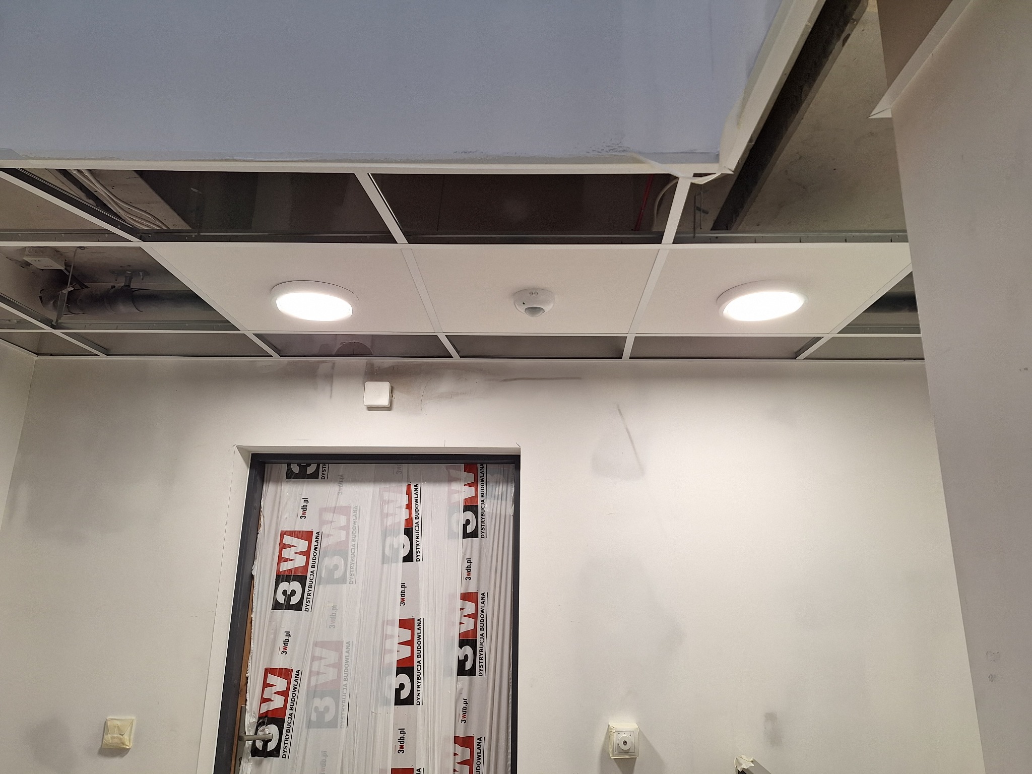 Sufit podwieszany z rastrem, oświetleniem LED i czujnikiem dymu, widoczne kable w przestrzeni nad sufitem, ściana z drzwiami zabezpieczonymi folią z nadrukiem.