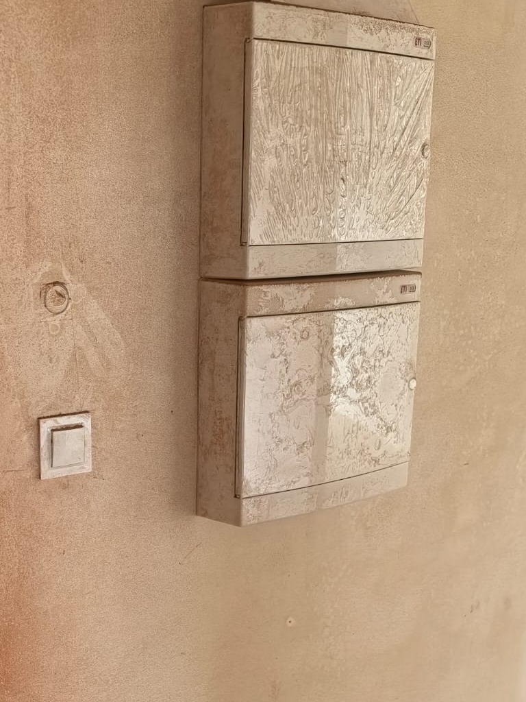 Dwa brudne, białe panele elektryczne ETI zamontowane jeden nad drugim na beżowej, teksturowanej ścianie obok kwadratowego włącznika światła.