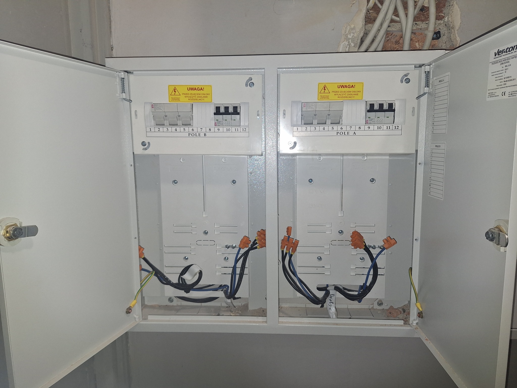 Otwarta, biała skrzynka rozdzielcza z bezpiecznikami i okablowaniem w trakcie montażu. Widoczne oznaczenia 'Pole A' i 'Pole B' oraz żółte naklejki ostrzegawcze.